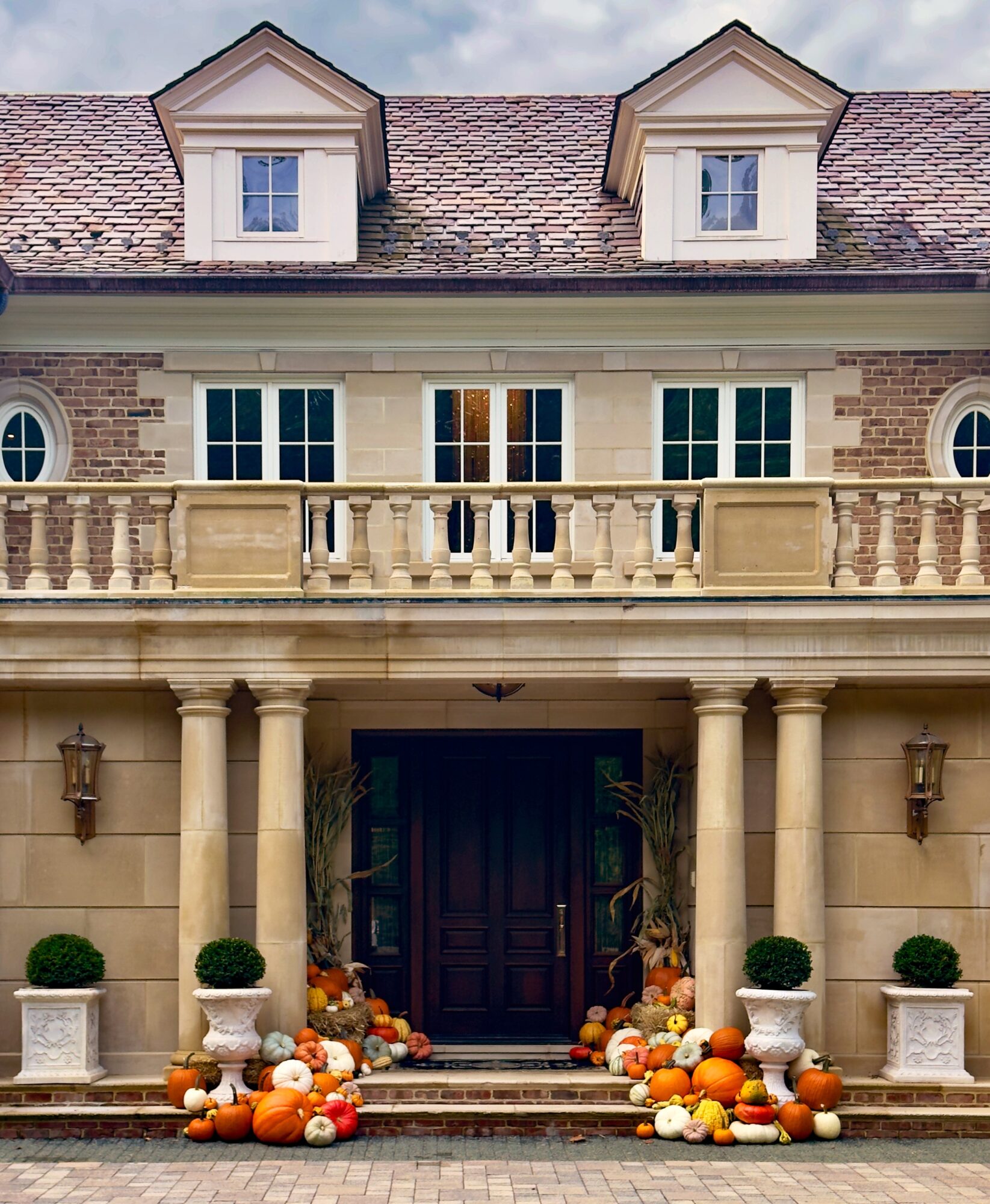 Front entrance of a large house with columns, pumpkins, and potted plants, under a cloudy sky.