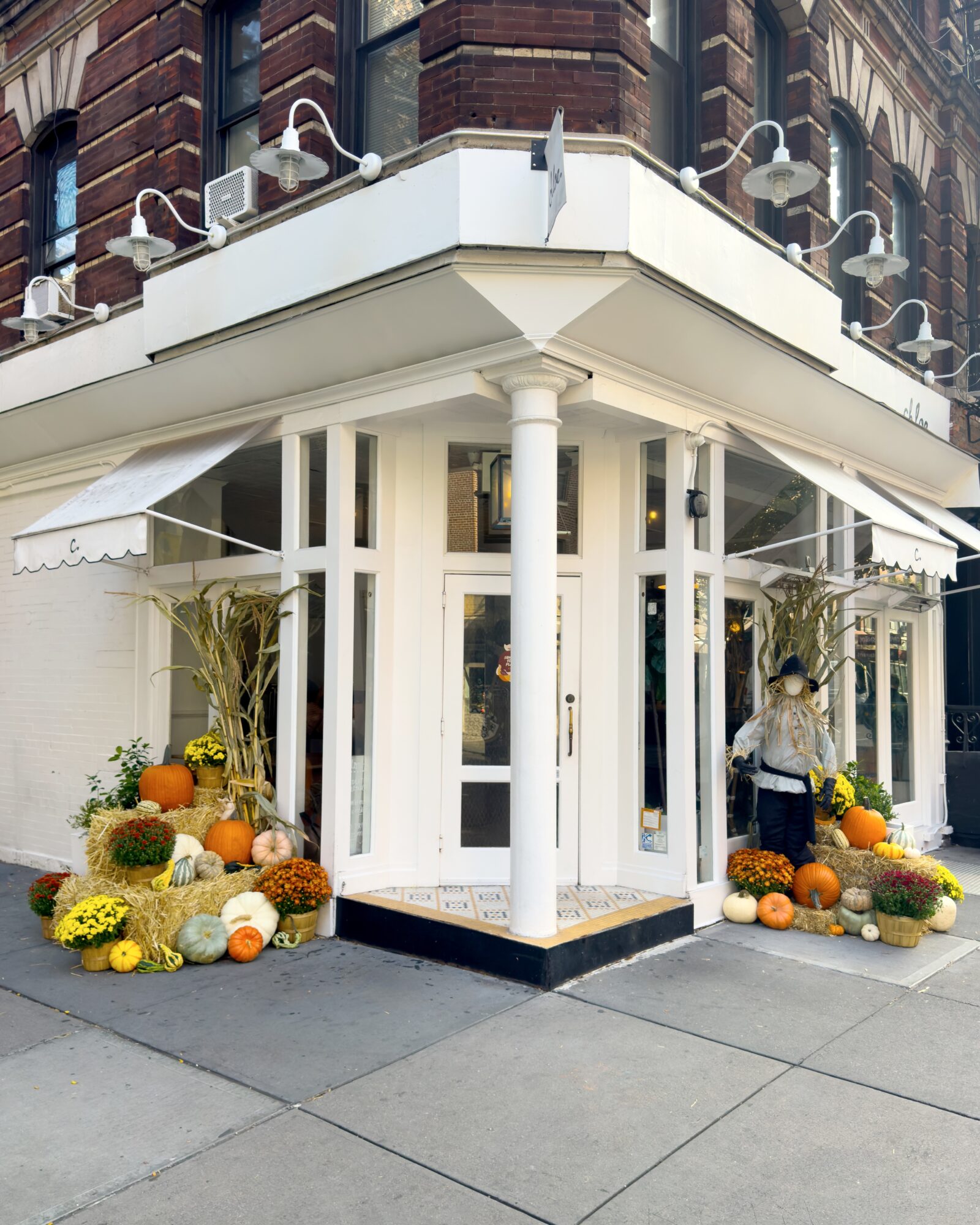 Corner storefront with white trim, decorated with pumpkins, gourds, and flowers outside, on a city sidewalk.