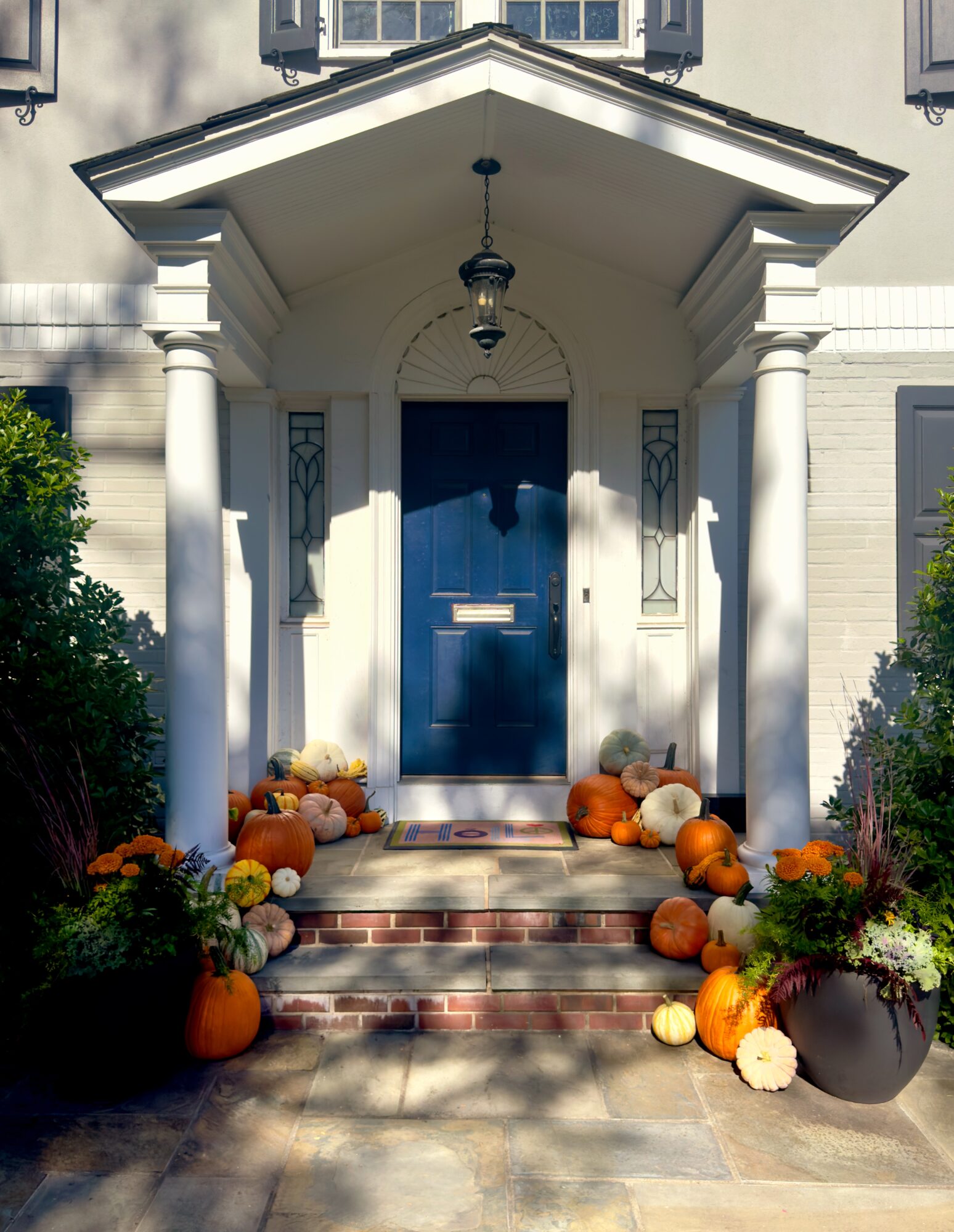 Front porch with blue door, white columns, and fall decorations including pumpkins and flowers, sunlight casting shadows.