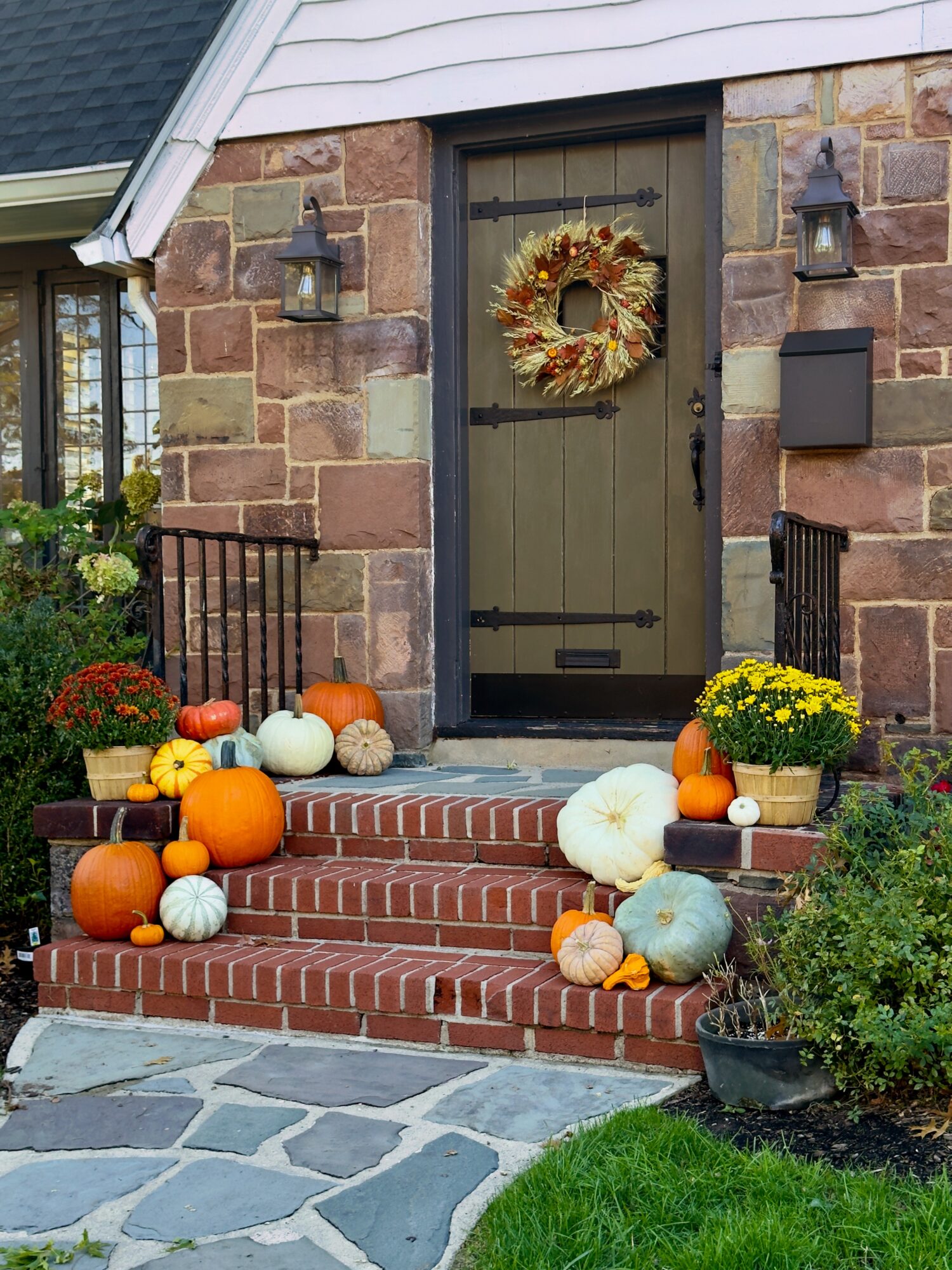 Front door decorated with pumpkins and a wreath, surrounded by potted plants and fall decorations on brick steps.