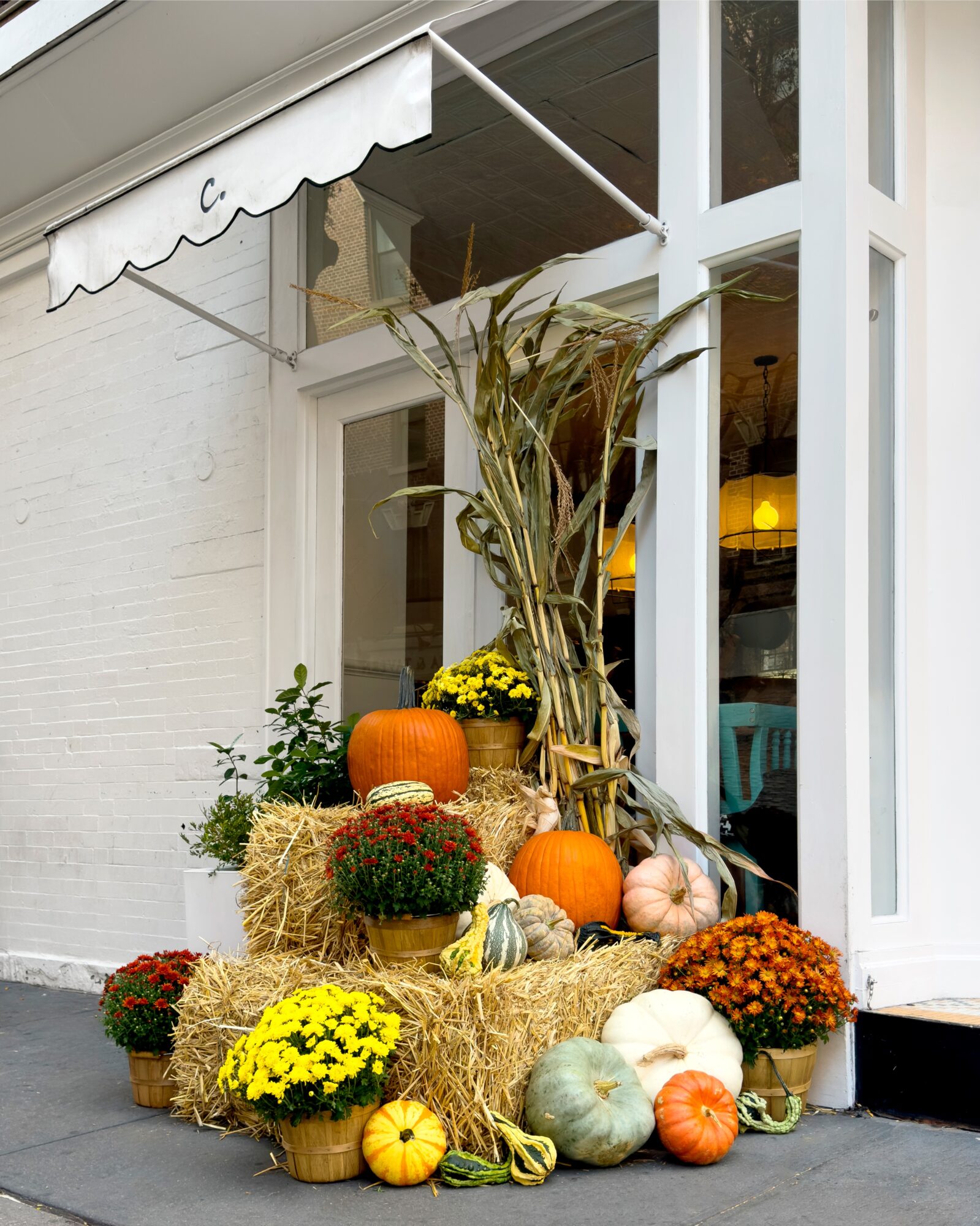 Fall display with pumpkins, gourds, hay bales, and mums outside a glass door with a white awning.