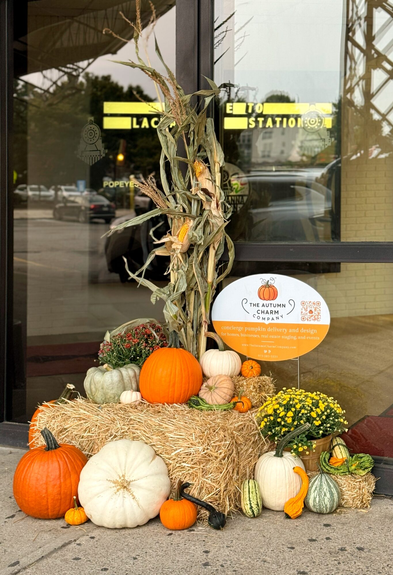 Fall display with pumpkins, gourds, hay, and flowers outside a building, with a sign and window in the background.