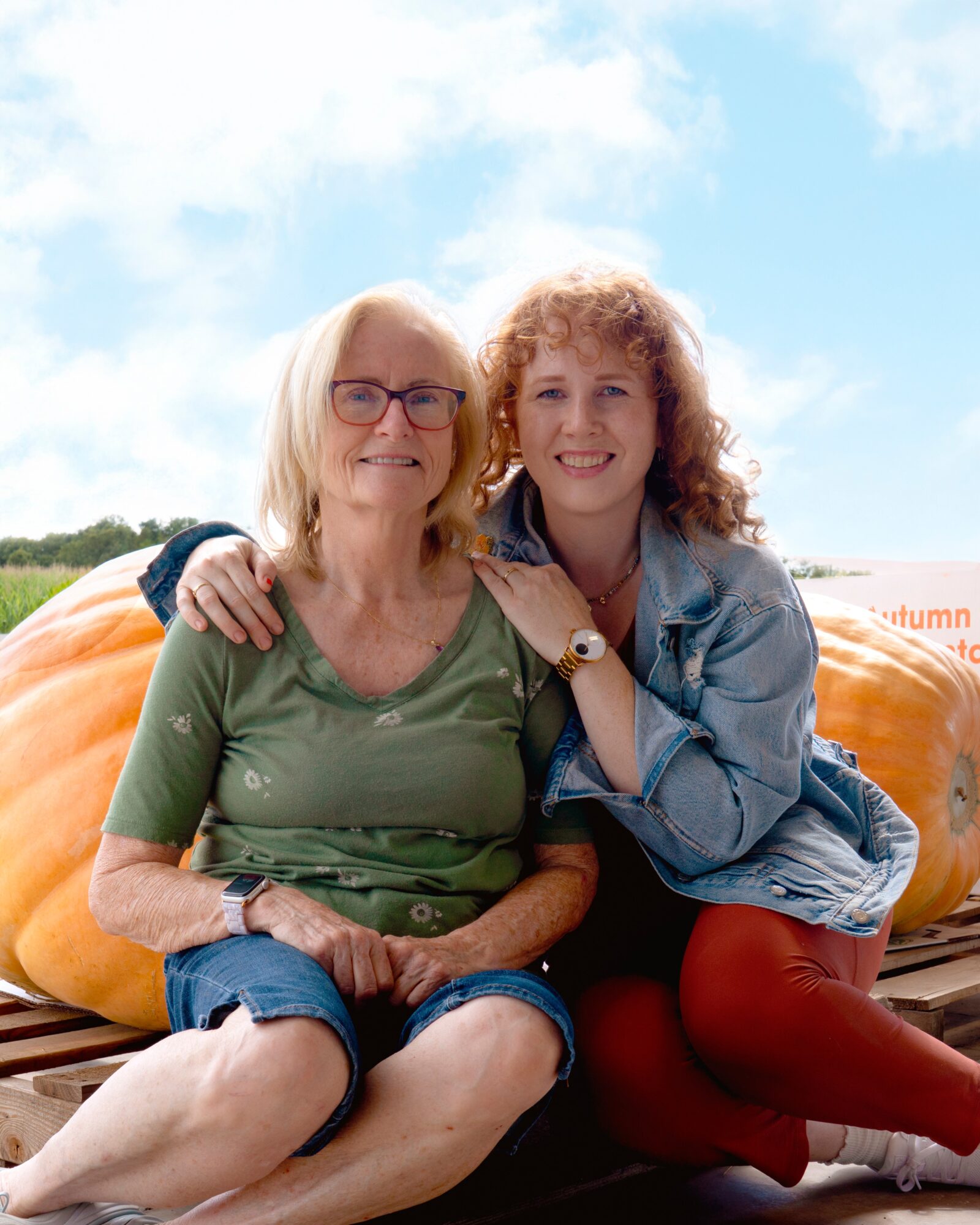 Two women sitting outdoors, smiling, with a large pumpkin behind them, one with glasses and the other with curly hair.