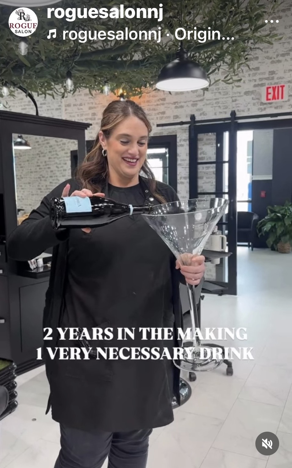 Woman in black apron pouring drink into a large martini glass in a modern bar, smiling, with white brick wall and plants in background.