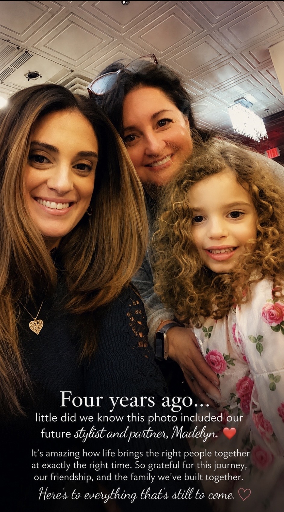 Three people smiling indoors, two women and a young girl with curly hair, close together, in a warmly lit room.