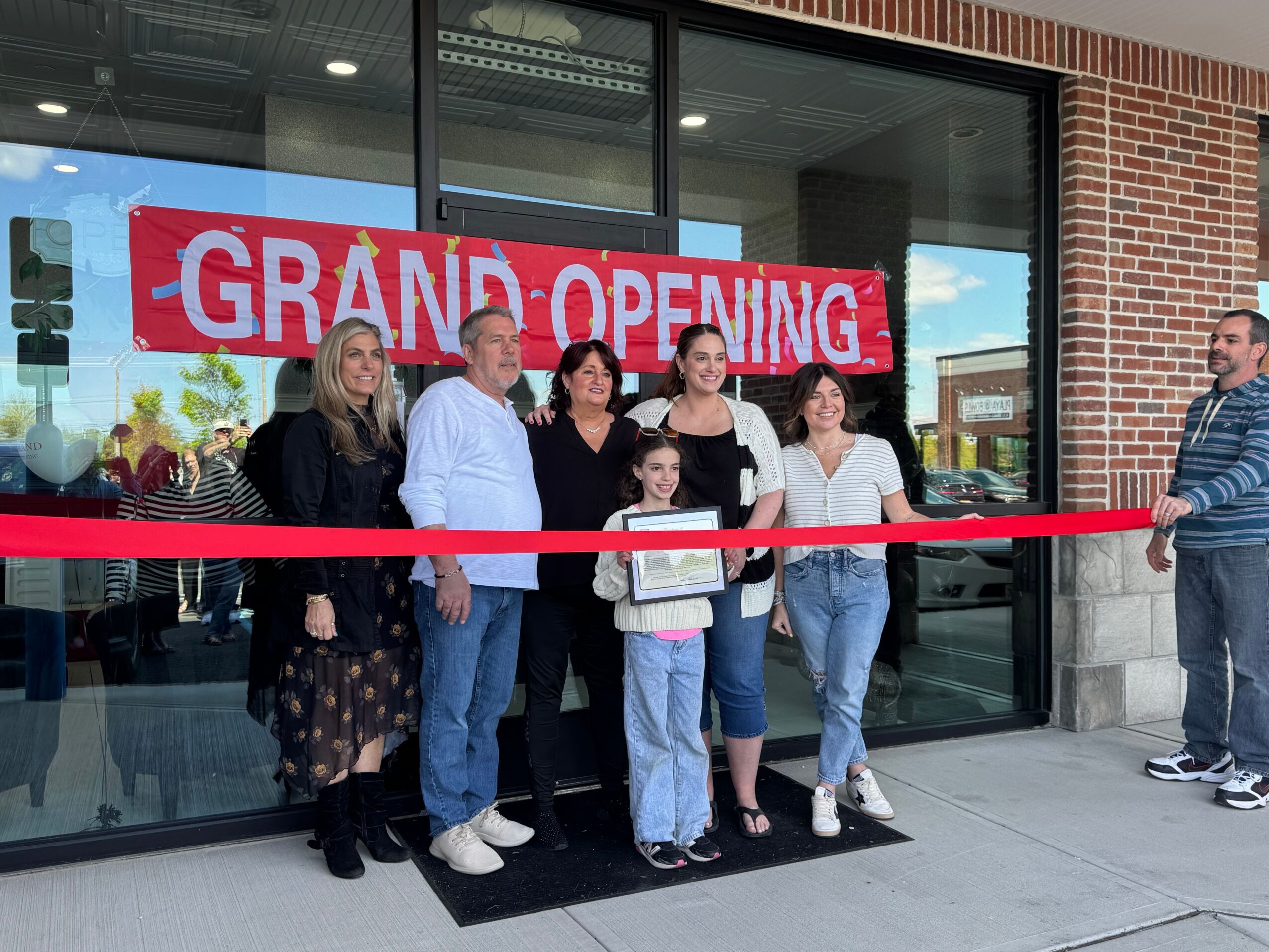 Group of people standing outside a building with a red and white 'GRAND OPENING' sign, some holding shopping bags.