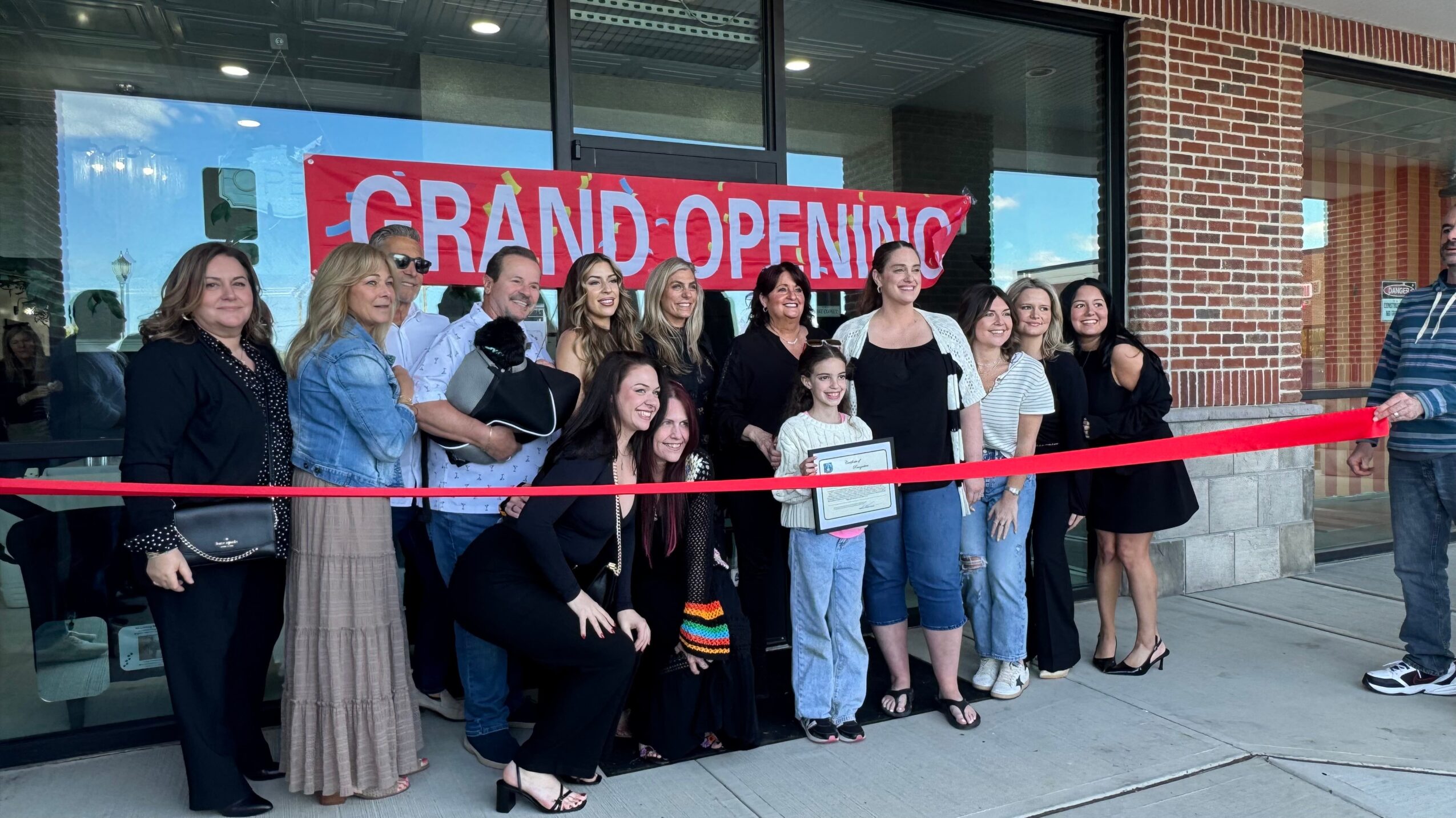 Group of people gathered outside a store with a red ribbon for grand opening, in front of a glass window with a red banner.