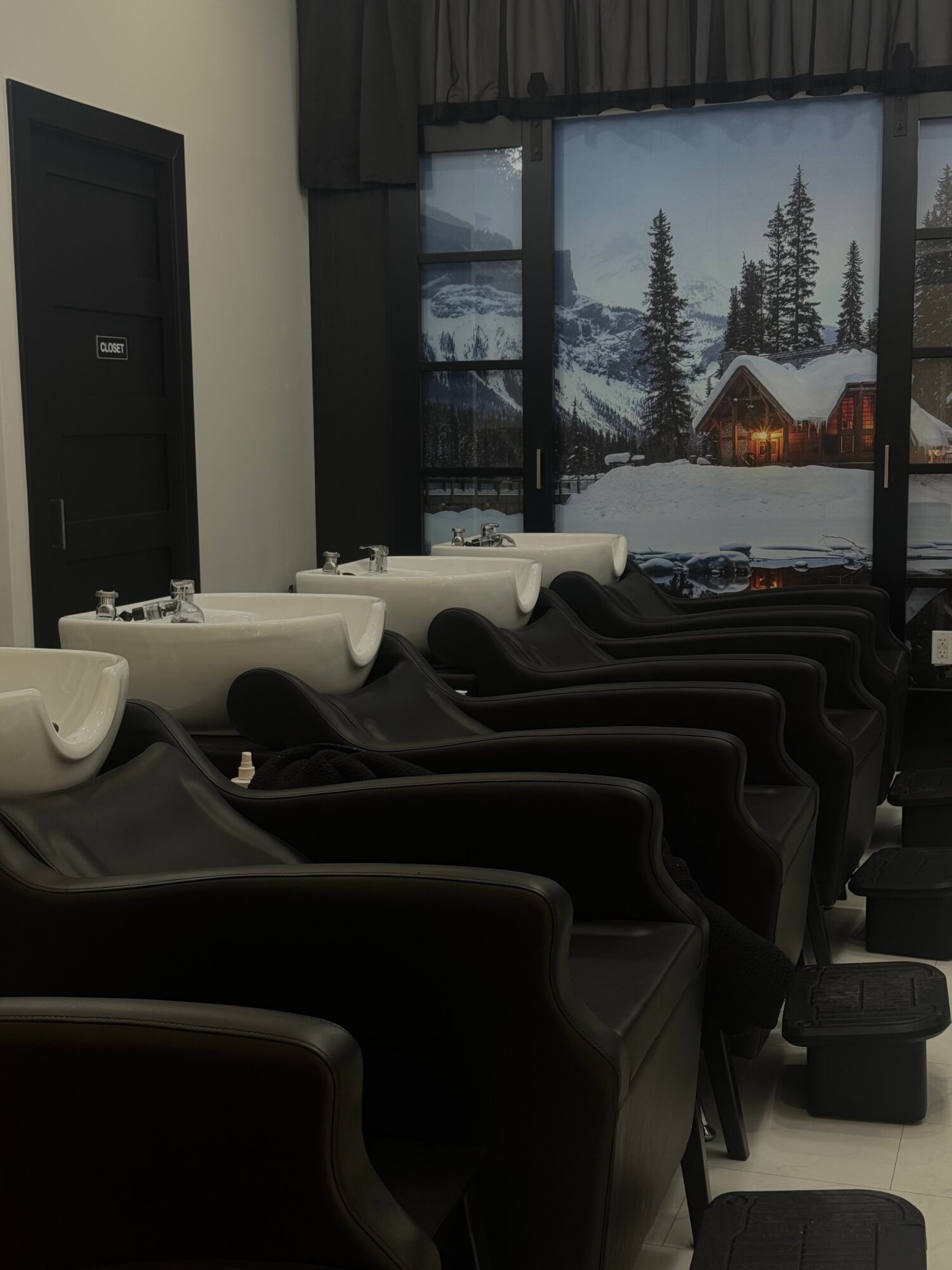 Salon with black chairs and white sinks, large window showing snowy landscape and trees outside.