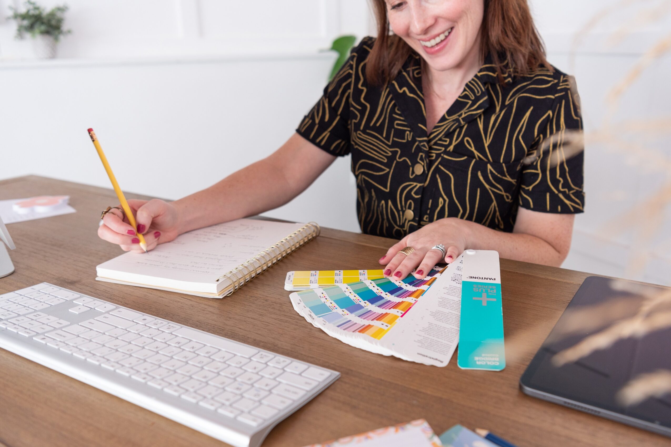 Woman sitting at desk with color swatches, notebook, keyboard, and tablet, smiling and writing with a pencil.