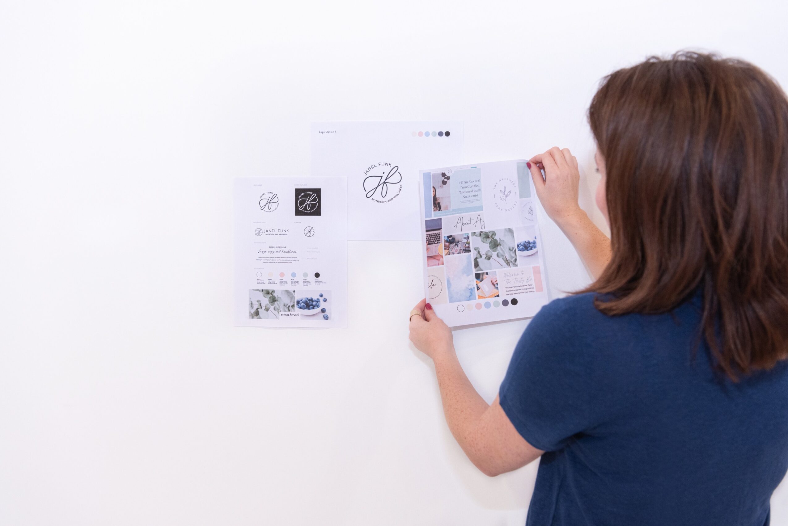 Person with brown hair in a blue shirt looks at a wall with papers and images attached, facing away from the camera.