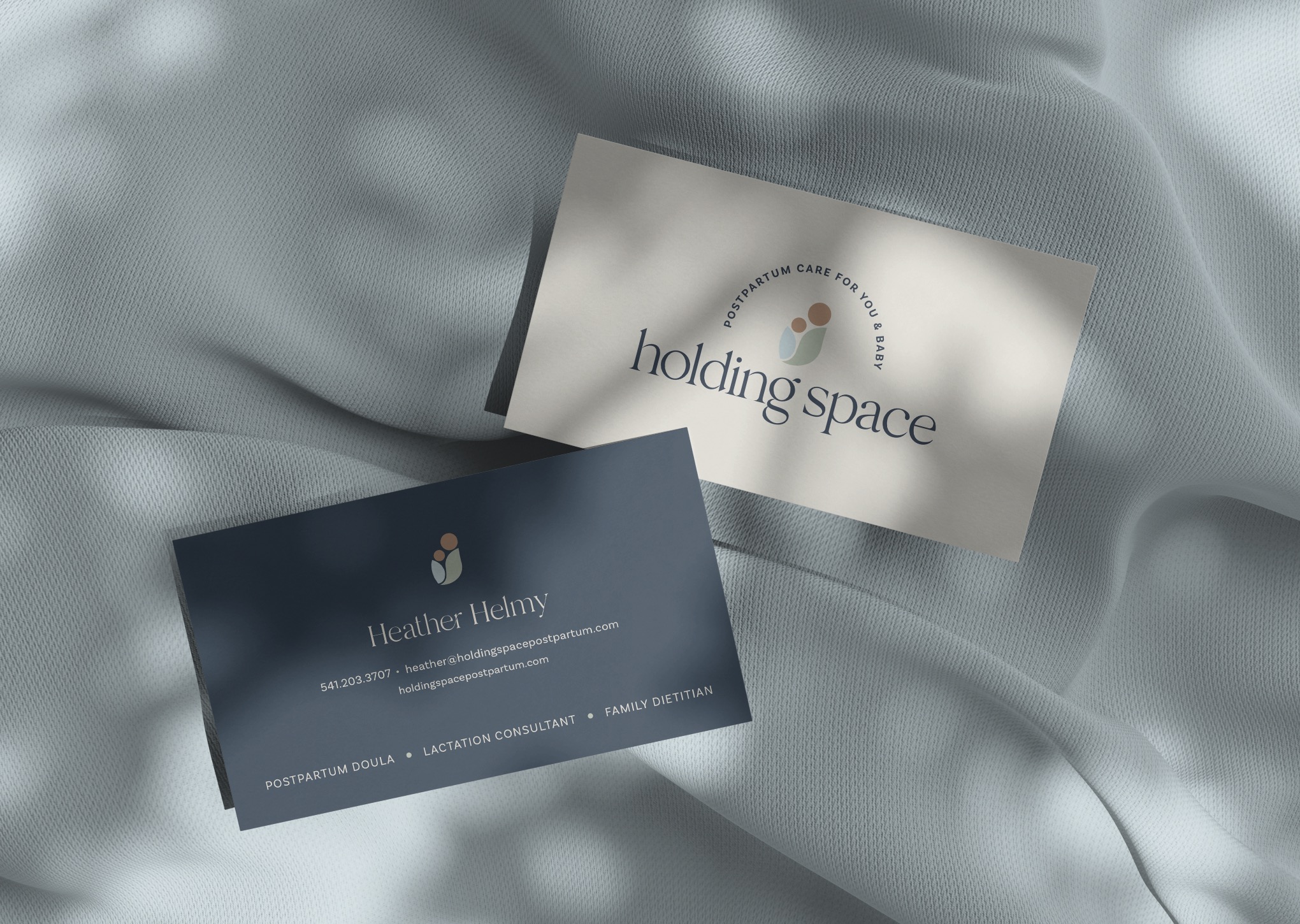 Two business cards on a soft, light-colored fabric surface, one with the text 'holding space' and the other with contact details.