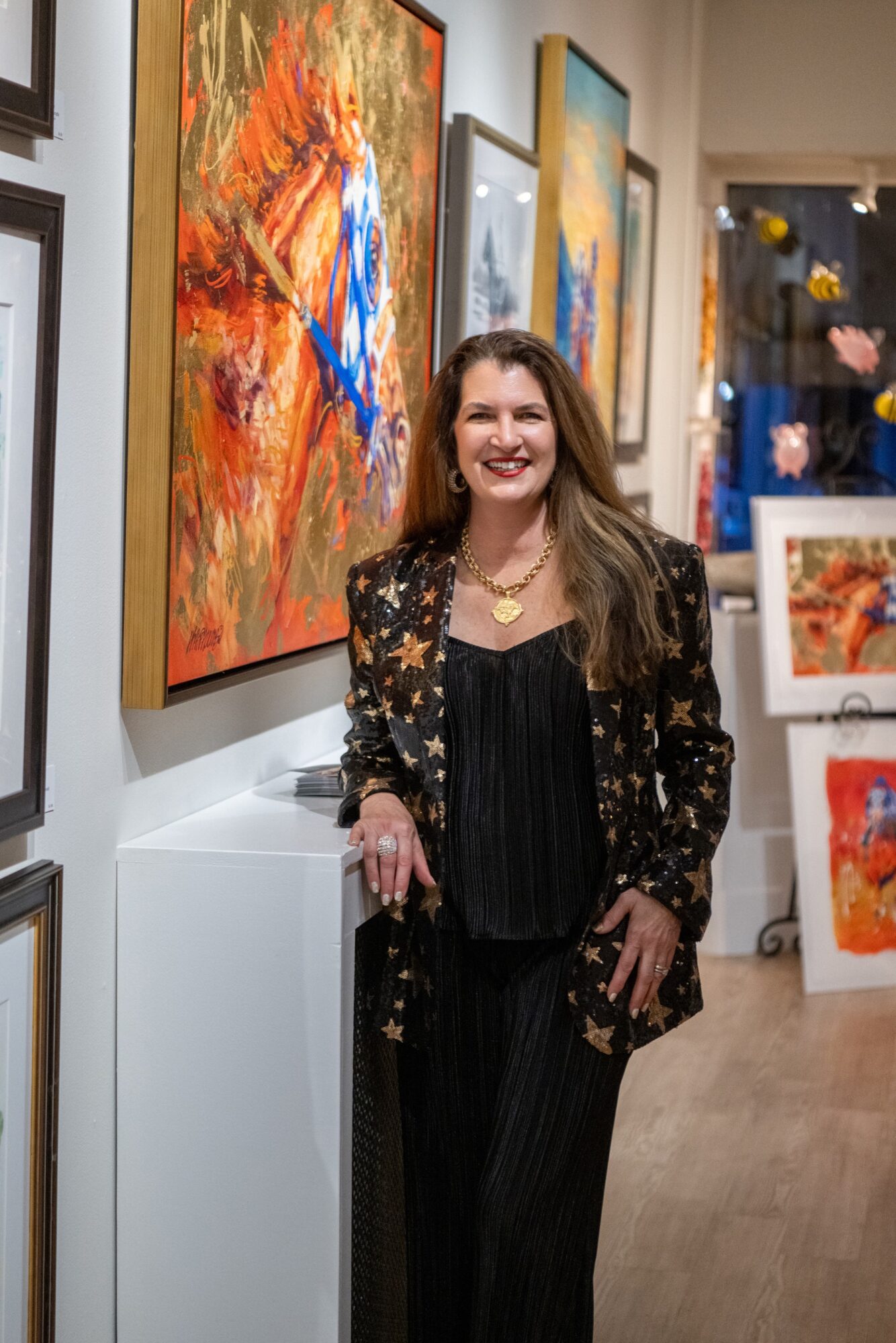 Woman with long hair smiling, standing in art gallery with colorful paintings, wearing a black outfit and star-patterned blazer.