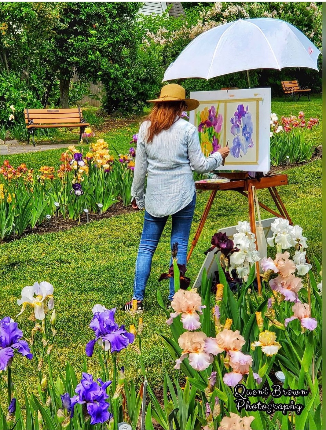 Woman painting outdoors in a garden with colorful flowers, under a large umbrella, with benches in the background.