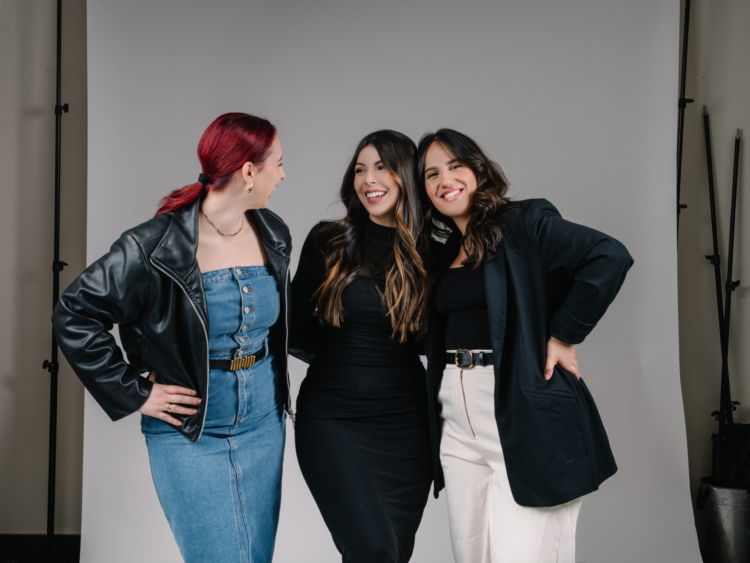 Three women standing together, smiling, in front of a plain background, with one woman wearing a leather jacket and jeans.