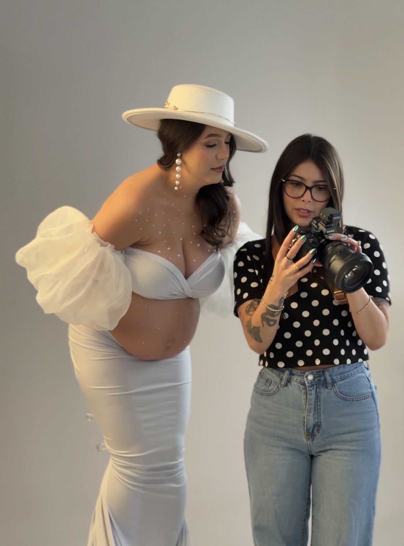 Two women taking a mirror selfie, one in a white outfit with a large hat, the other in a polka dot top and glasses.