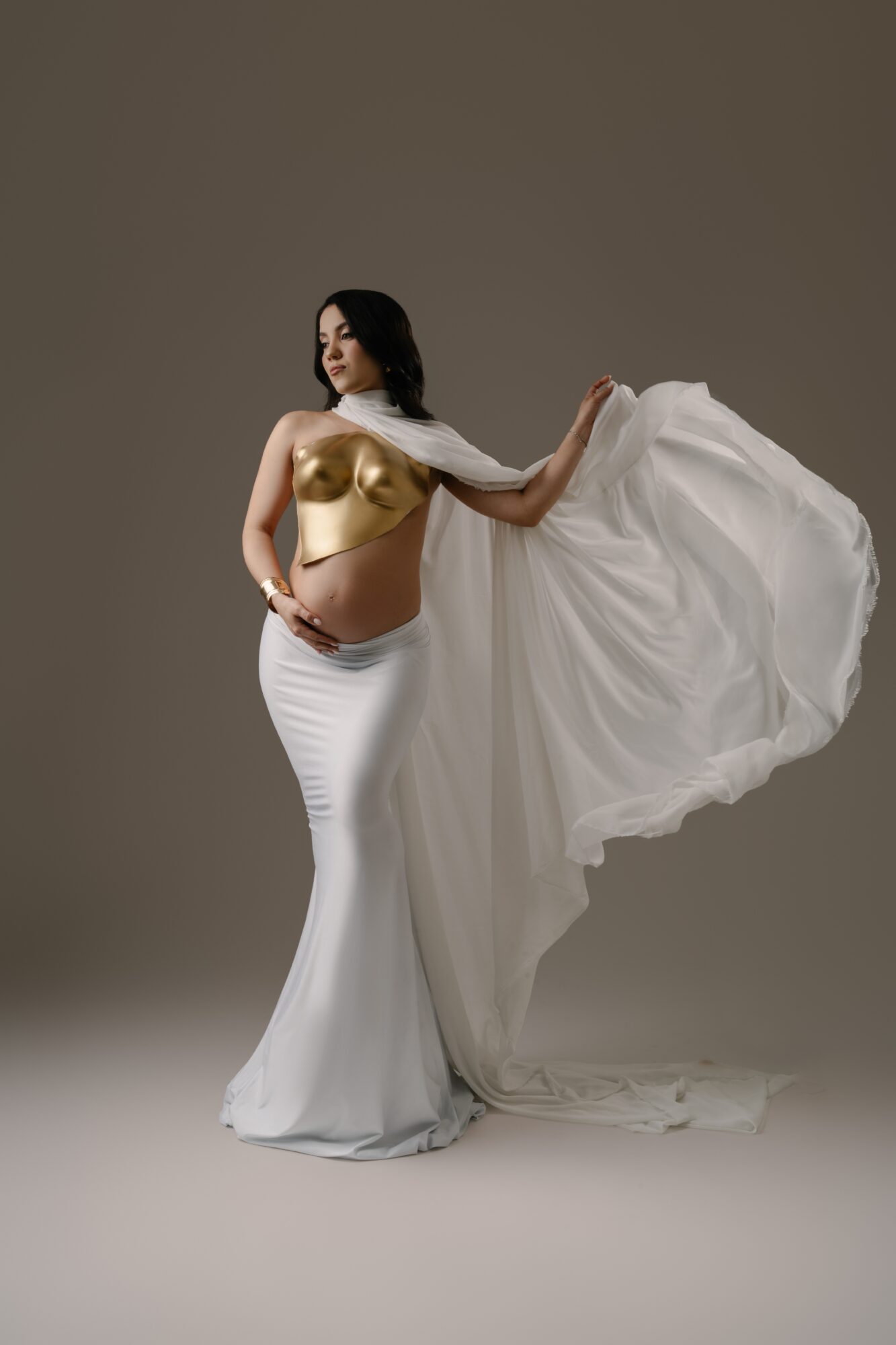 Woman in a white skirt and gold top holding a flowing white fabric, standing against a plain background.