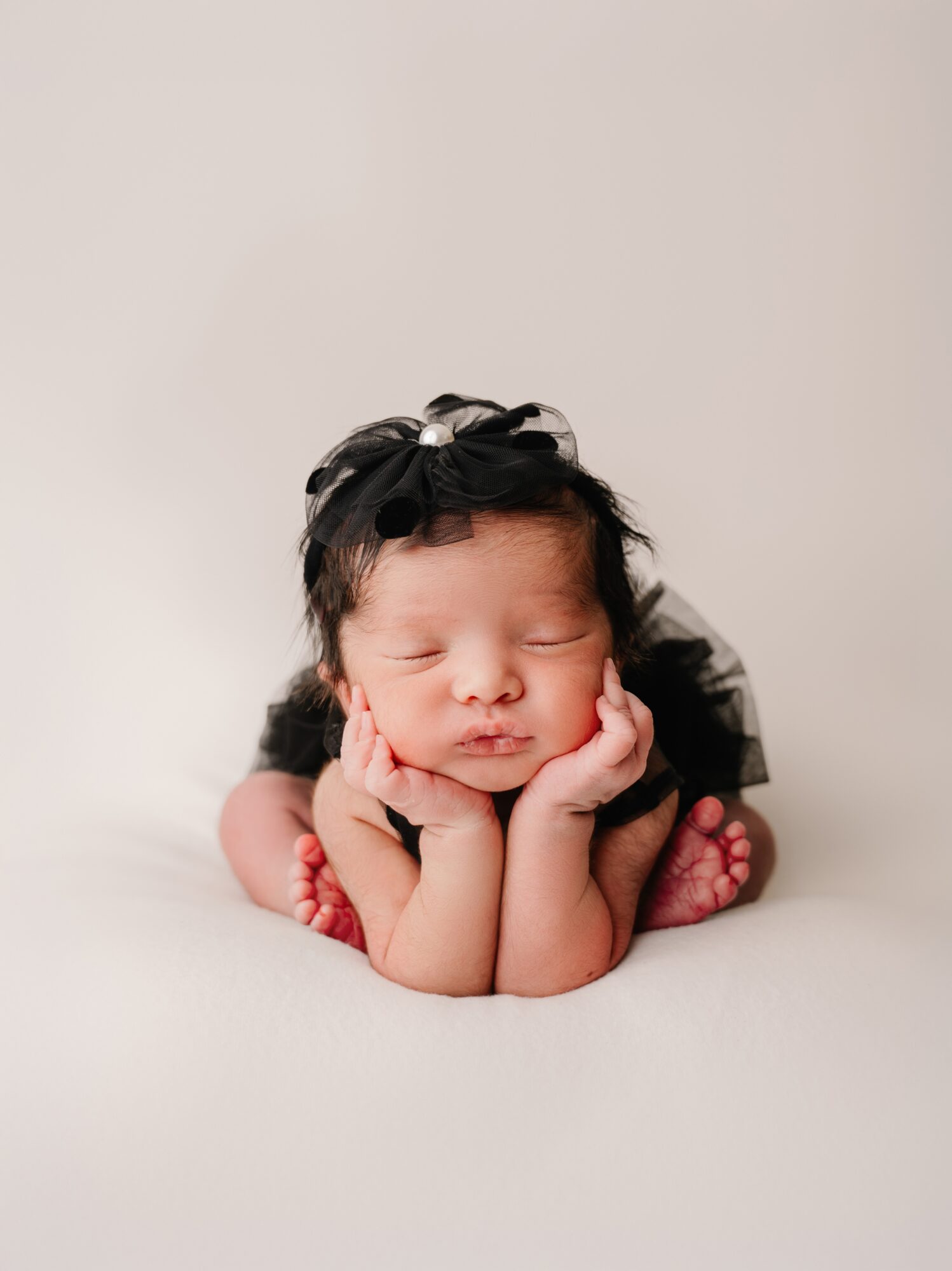 Young baby with a black bow headband, resting chin on hands, lying on a soft surface, with a neutral background.