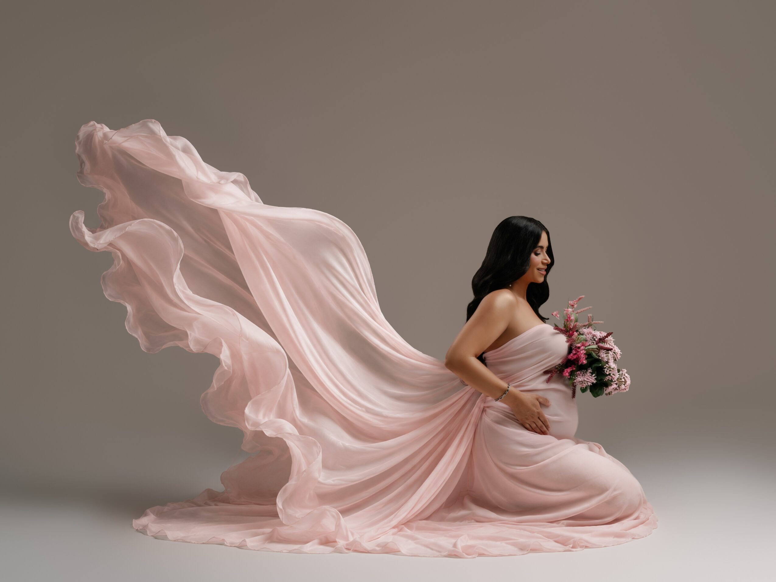 Woman in a flowing pink dress holding a bouquet, kneeling on the ground, with a long train flowing behind her.