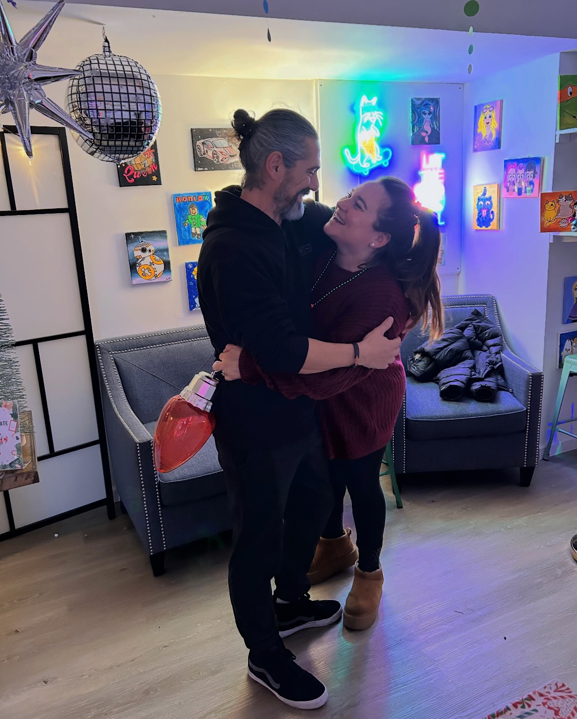 Two people dancing closely in a colorful room with artwork and neon lights on the wall.