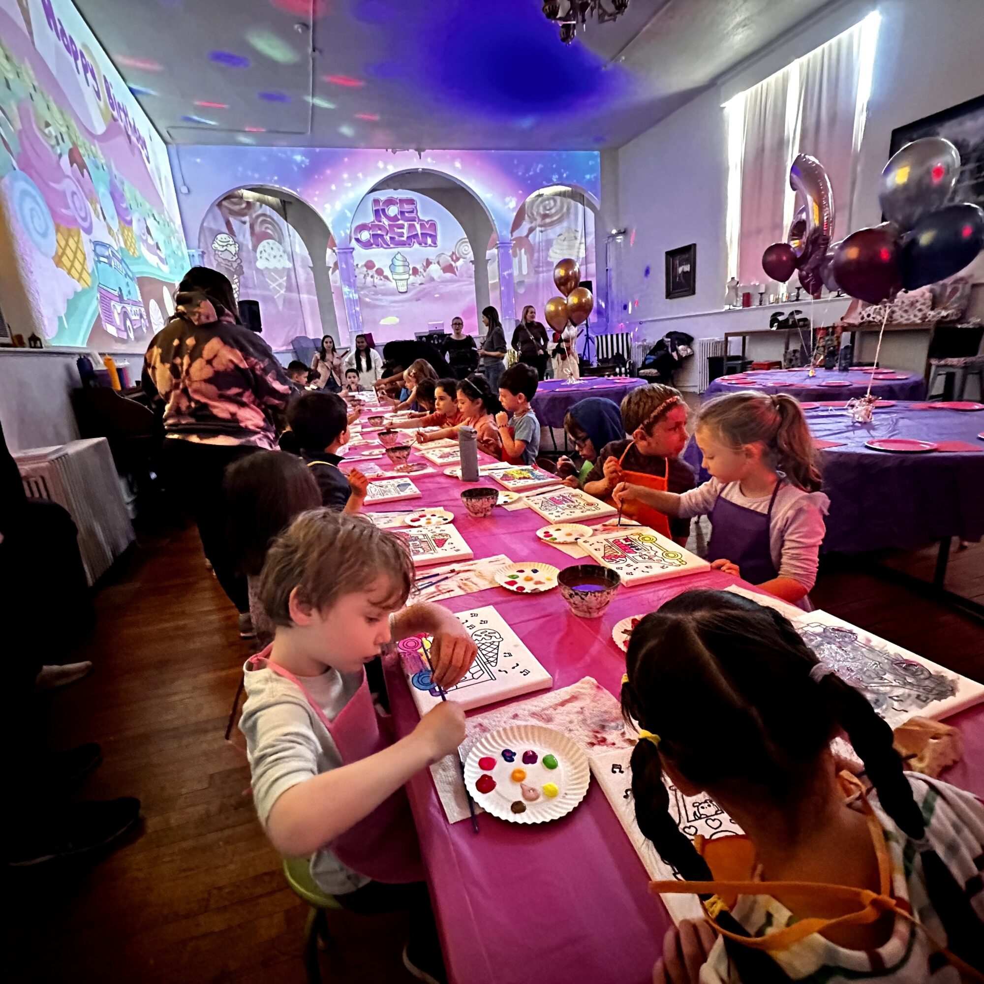 Children sit at a long table with pink tablecloth, engaging in arts and crafts during a party with colorful decorations and balloons.