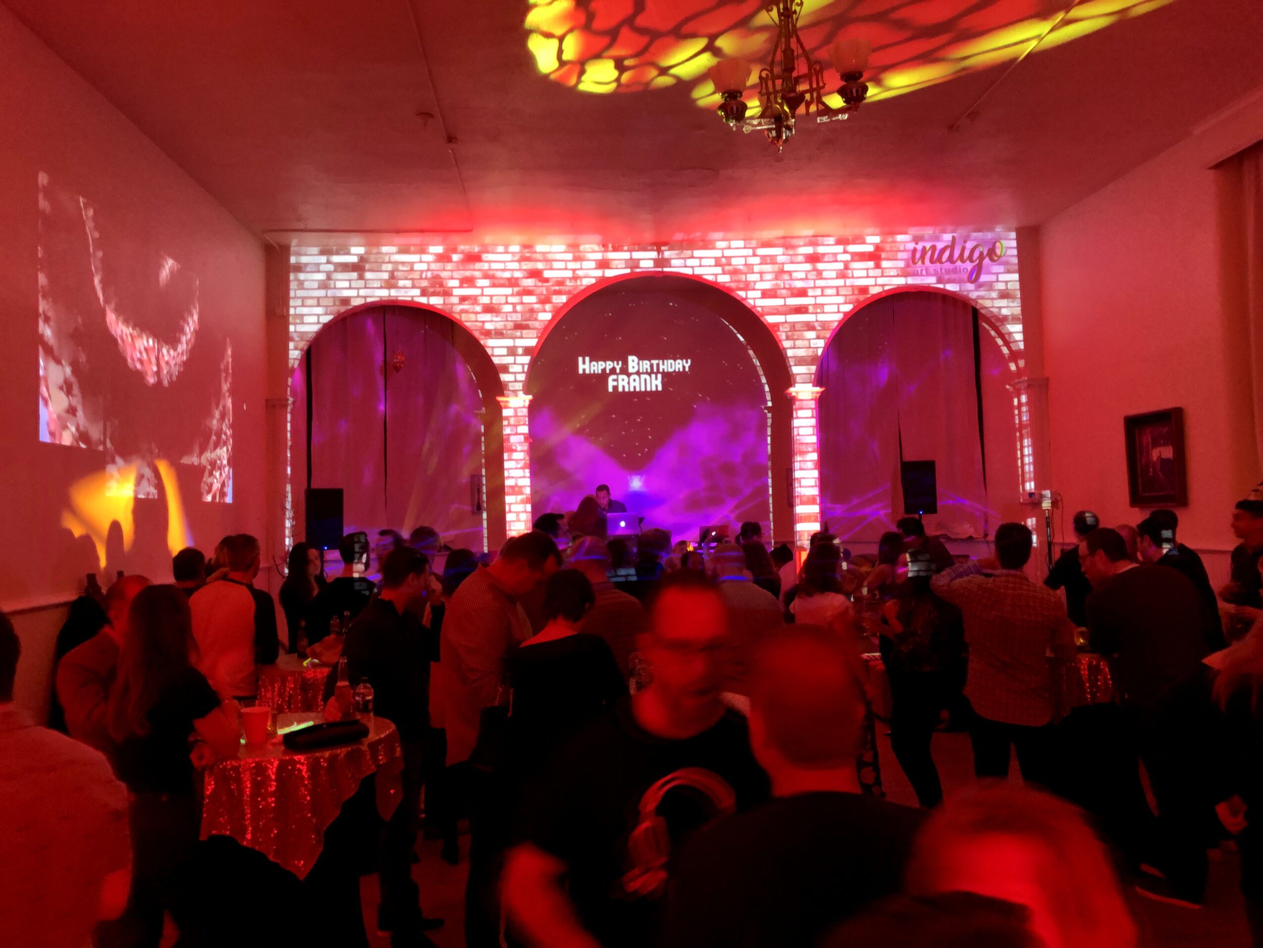 Indoor event with a stage, audience, and red lighting, featuring three arches and a chandelier, celebrating a birthday.
