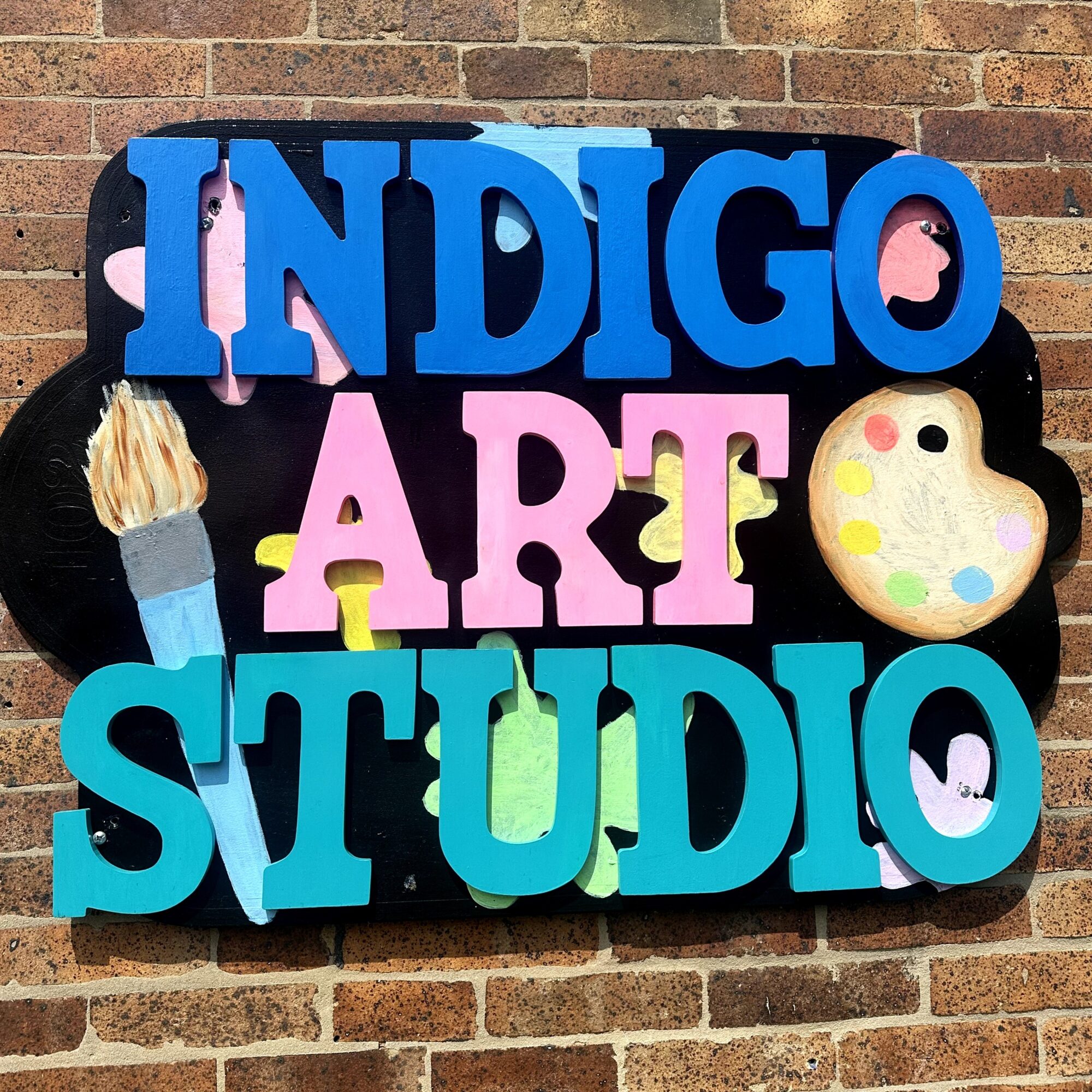 Colorful sign with words 'INDIGO ART STUDIO' on brick wall, decorated with paintbrush and palette illustrations.