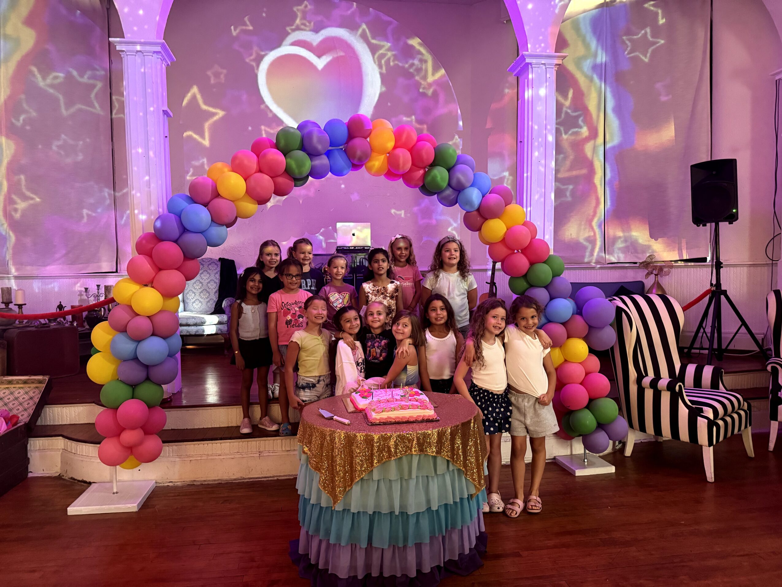 Group of children standing under a colorful balloon arch at a celebration with a decorated cake on a table.
