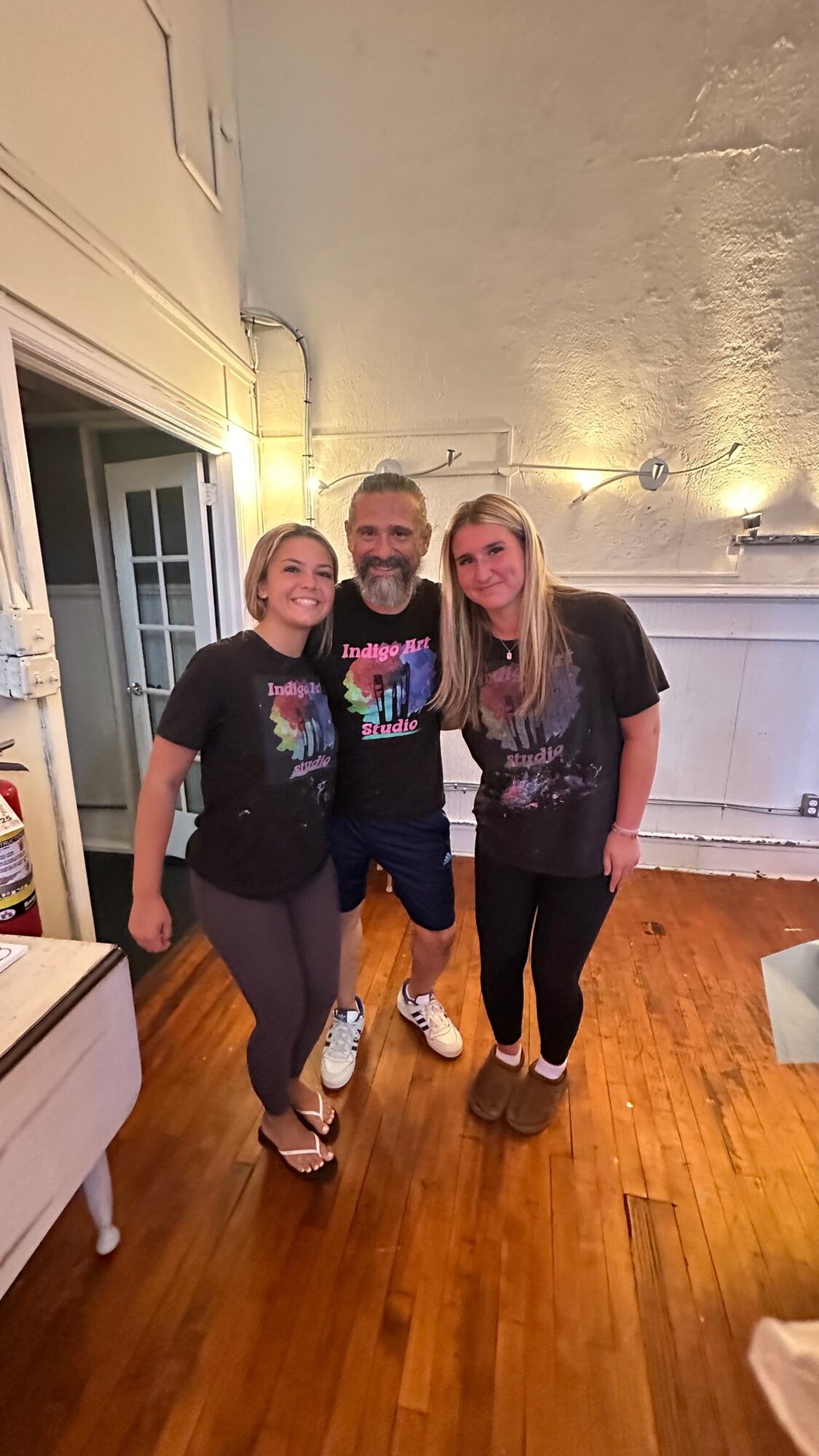 Three people standing indoors, smiling, with a wooden floor and white walls, two women and one man in the center.