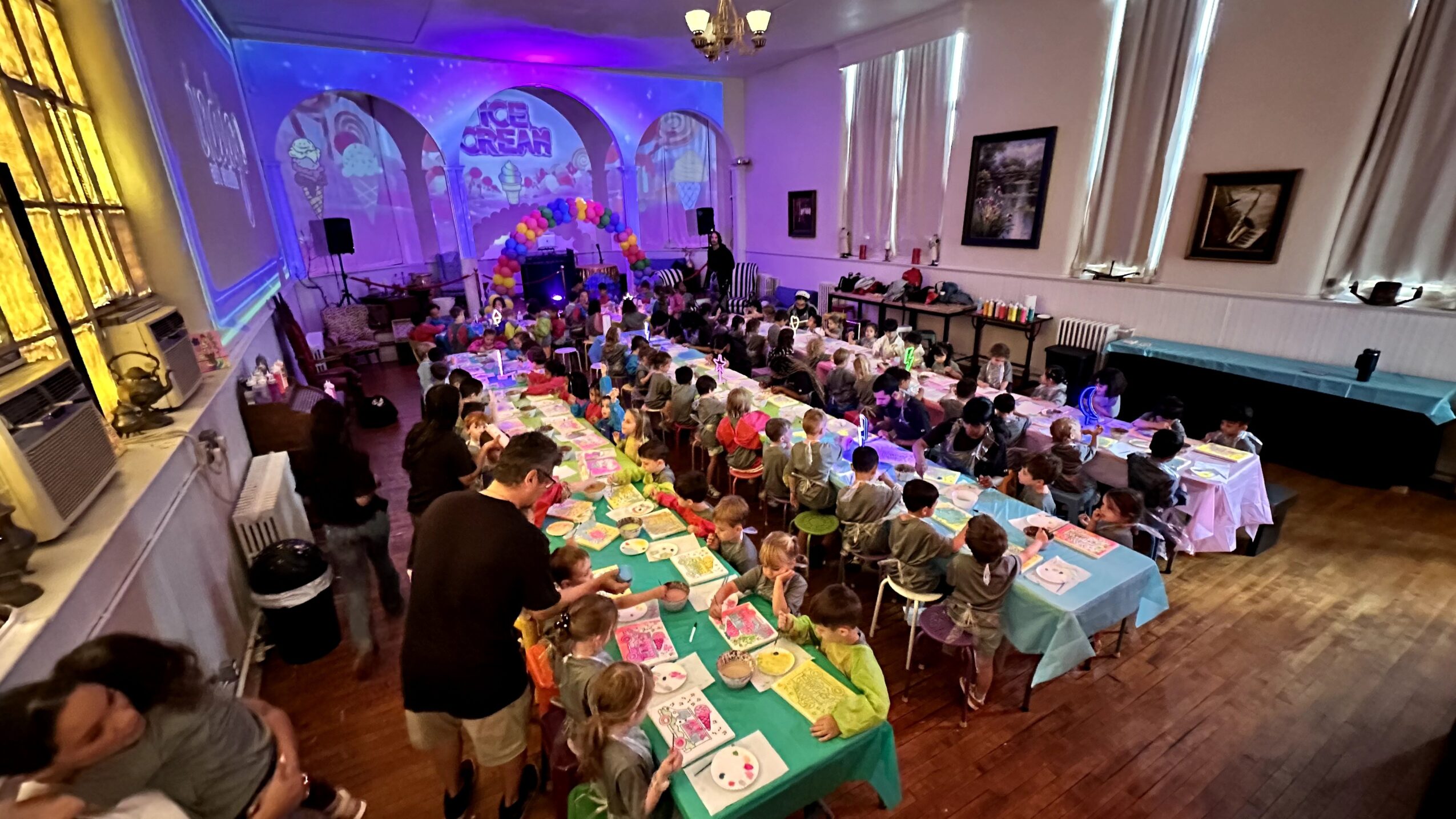 Large banquet hall filled with long tables of children and adults, colorful decorations, and a stage with blue lighting.
