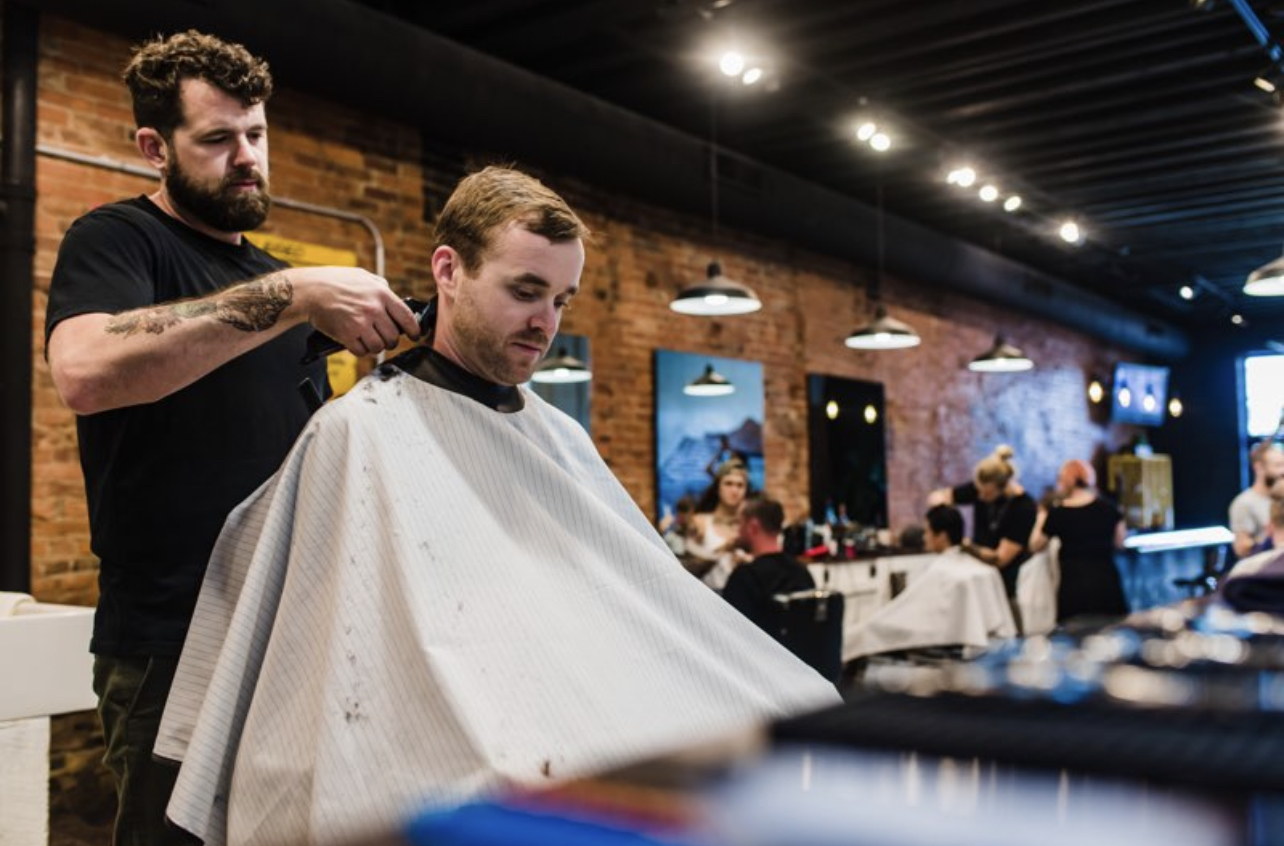 Barber cutting a man's hair in a salon with brick walls and hanging lights, customers seated at tables in background.