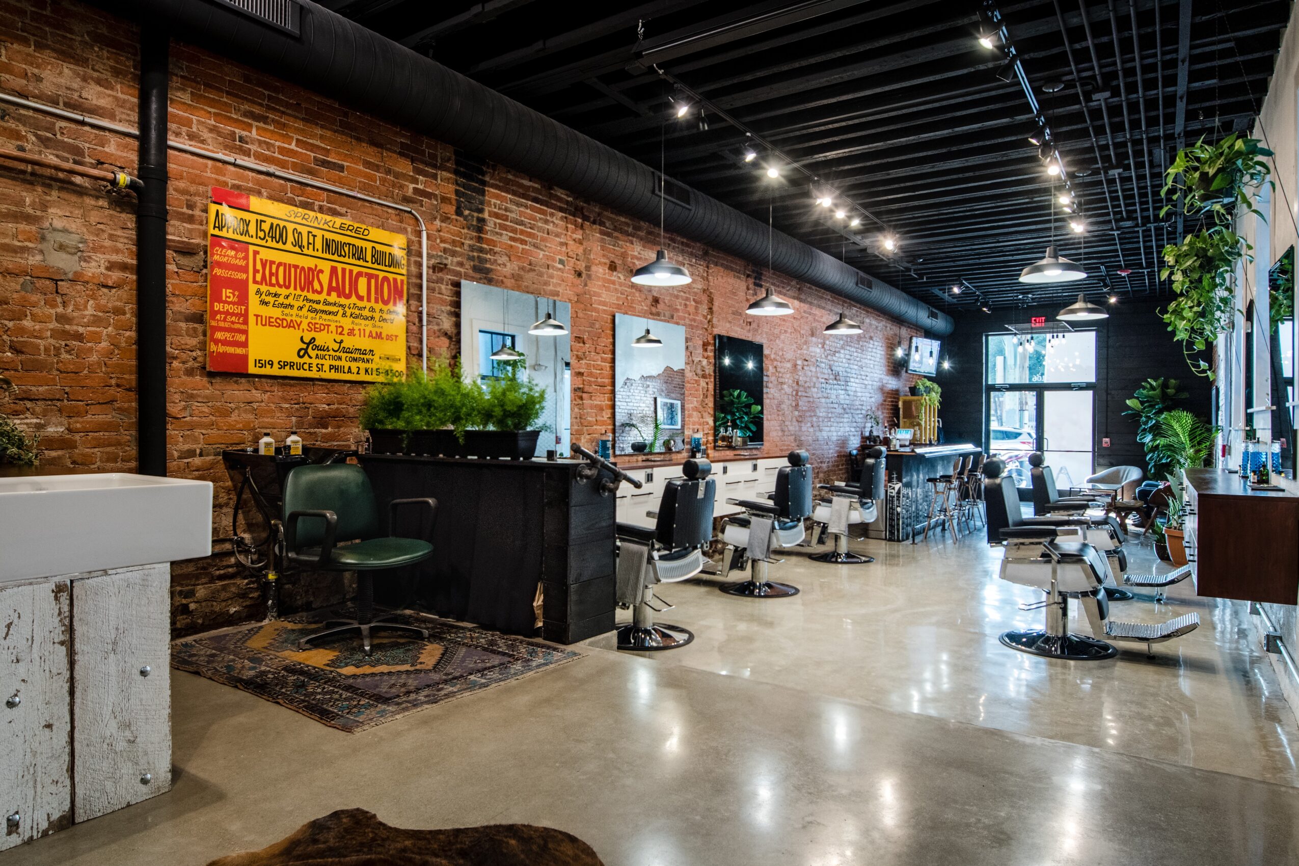 Modern hair salon with chairs, mirrors, and large windows, brick walls, and plants, illuminated by ceiling lights.