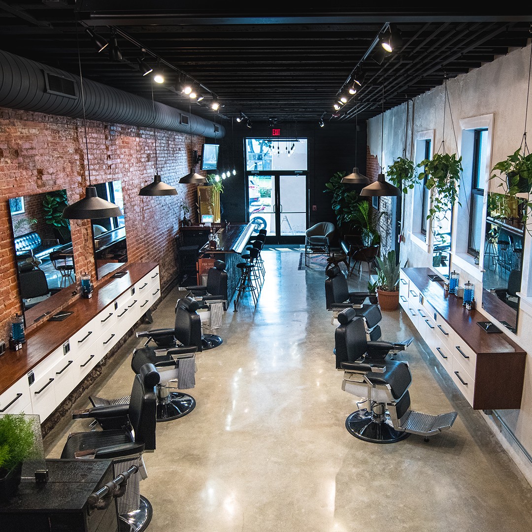 Hair salon interior with chairs, mirrors, plants, and large windows, illuminated by ceiling lights and natural light.