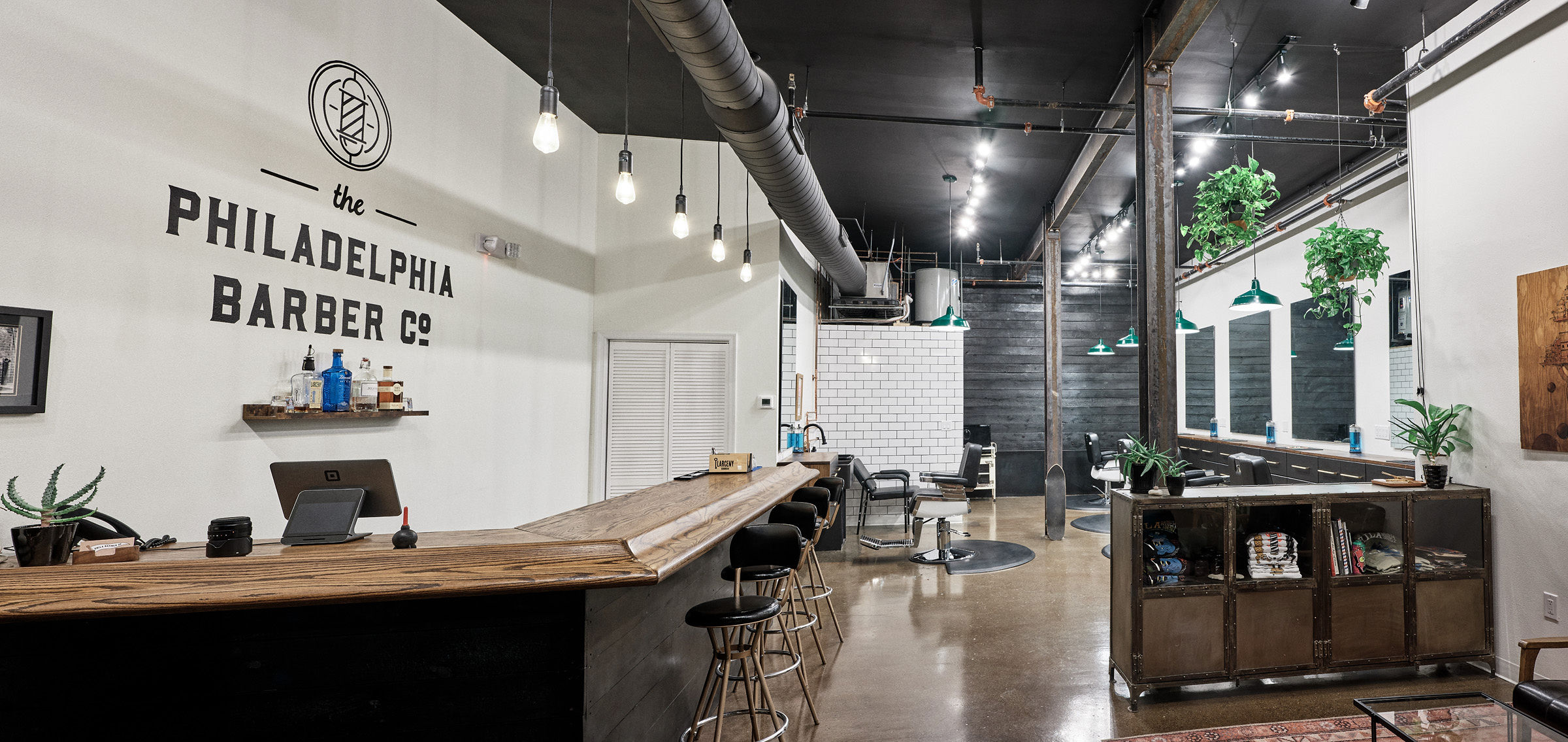 Interior of a barber shop with a wooden counter, chairs, and large windows, modern decor, and plants. Word sign reads 'Philadelphia Barber Co.'
