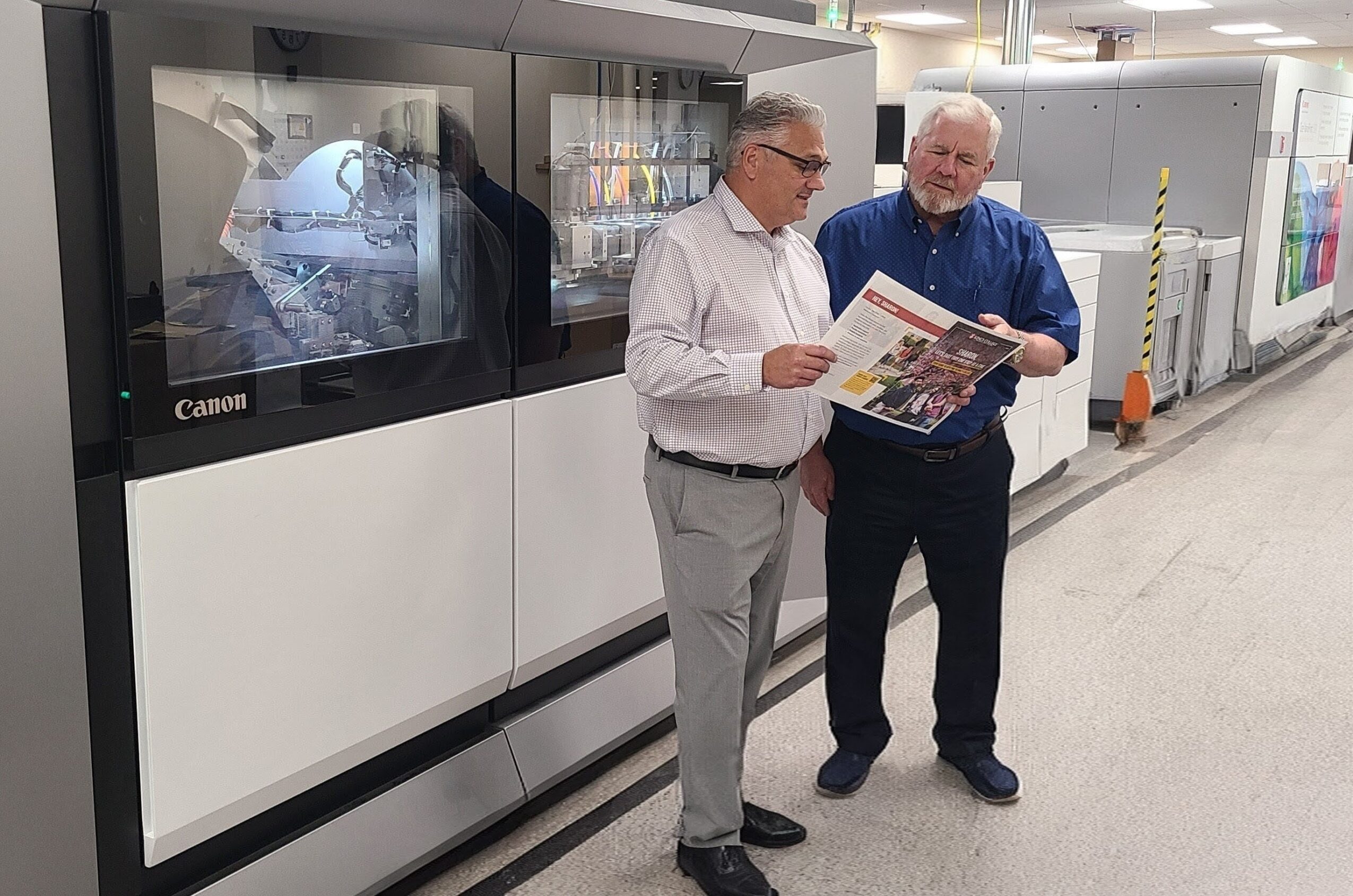 Two men stand in a hallway near a large machine, looking at a magazine together, with a bright, modern background.