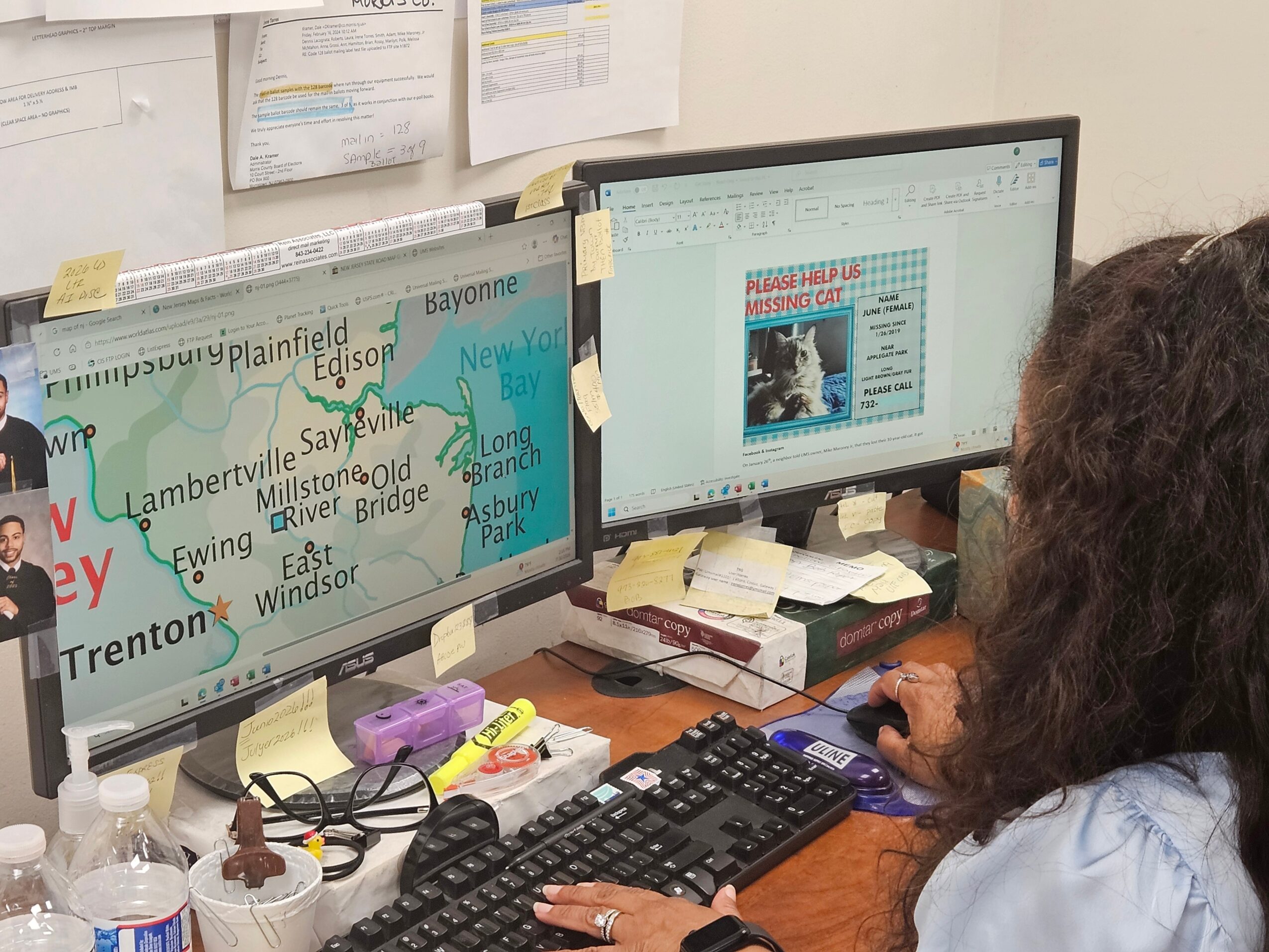 Two computer monitors on a desk with sticky notes, papers, and a person working on a keyboard, with a map on one screen.