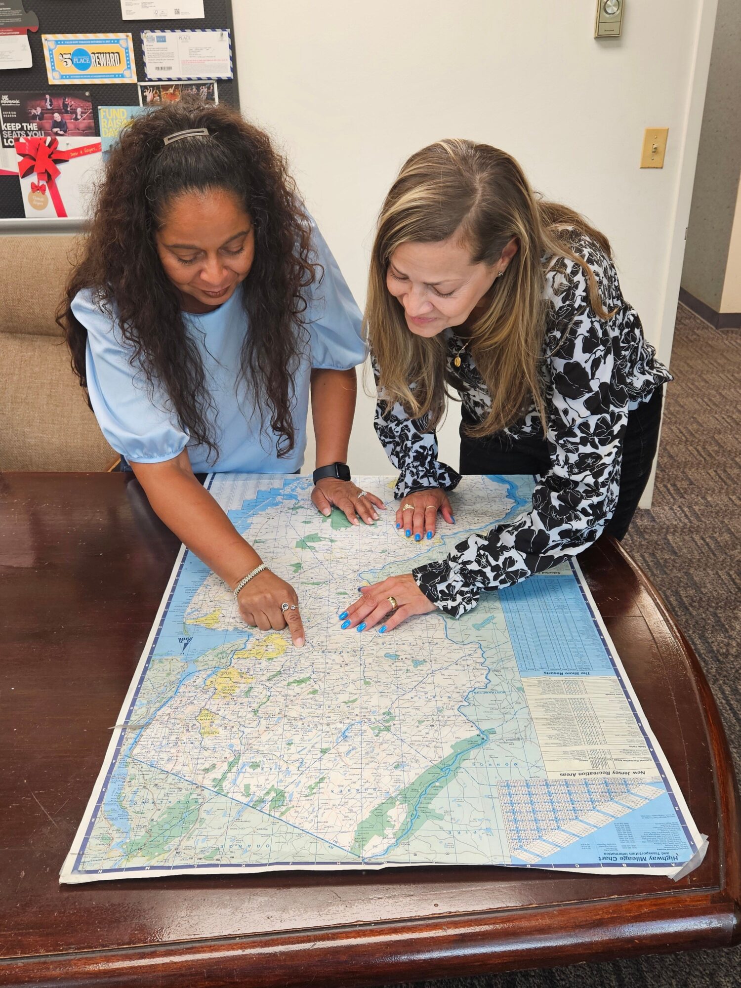 Two women look at a large map on a wooden table, one pointing at the map, in a room with a carpeted floor.