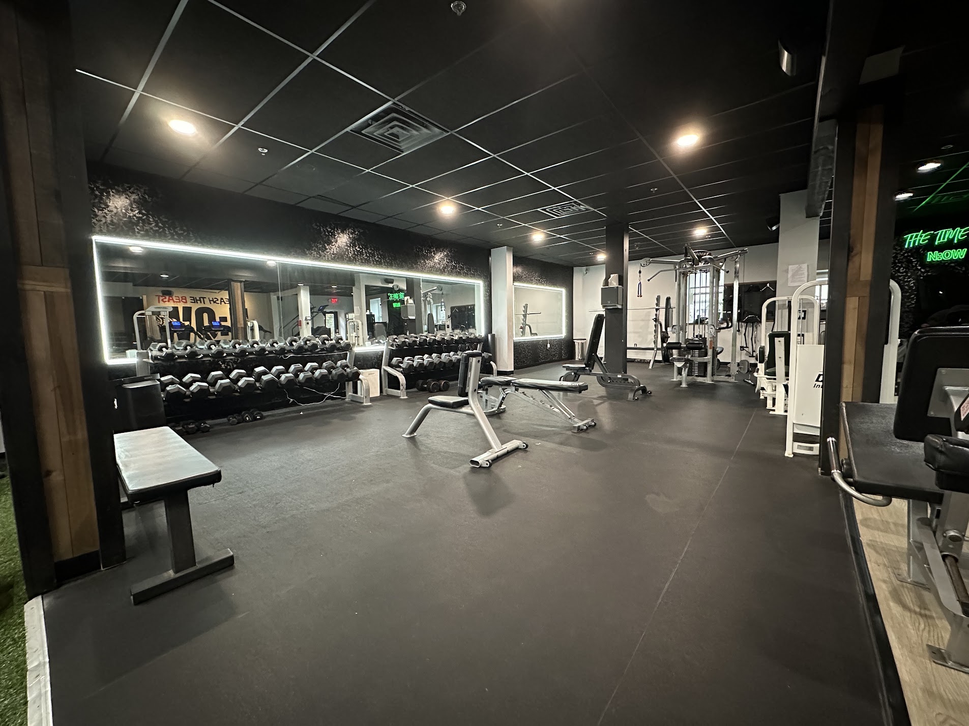 Empty gym with weight benches, dumbbells, and exercise machines, illuminated by ceiling lights, with a black ceiling and floor.
