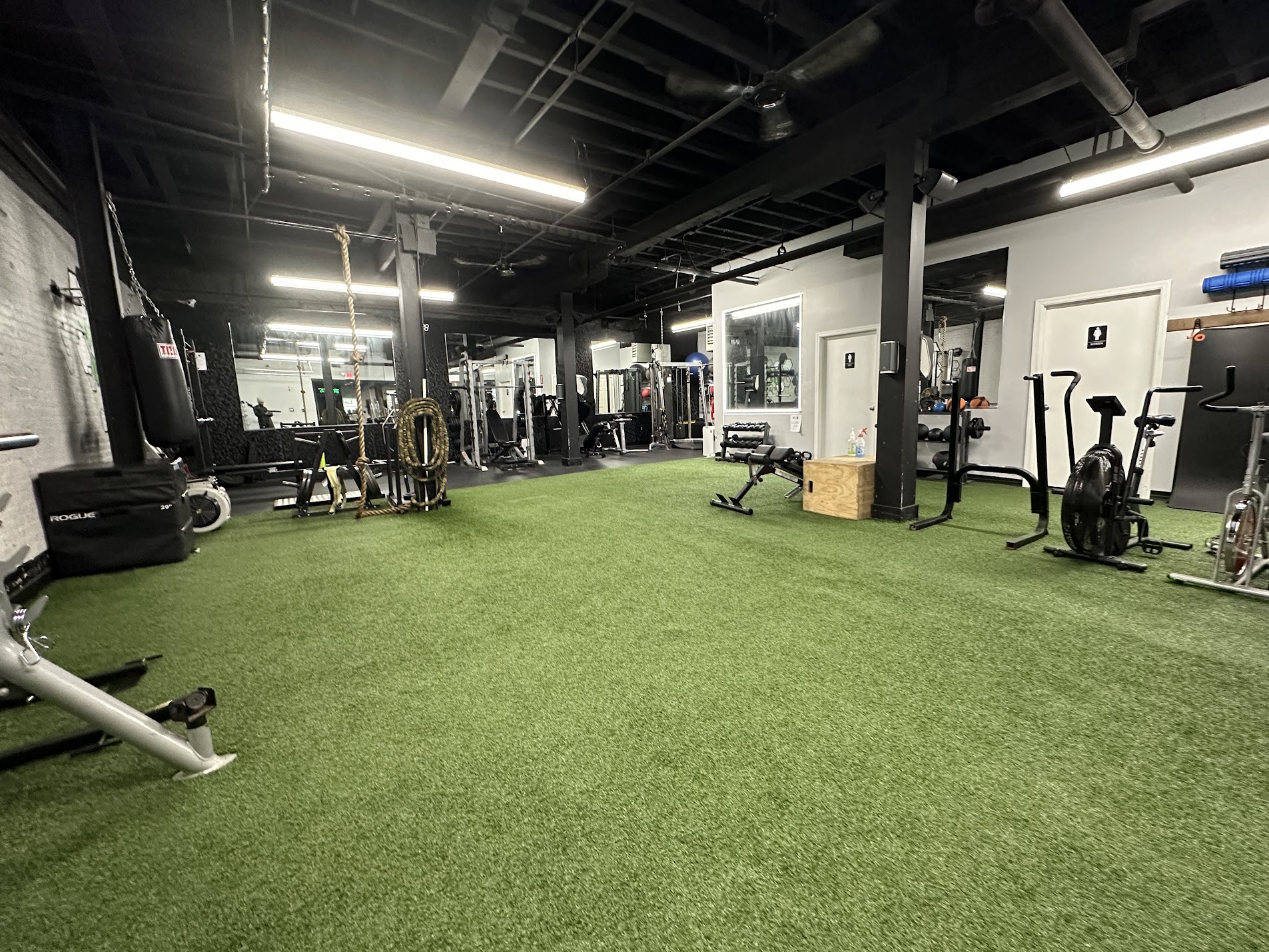 Empty gym with artificial grass flooring, exercise equipment, and large windows, under a black ceiling with lighting fixtures.