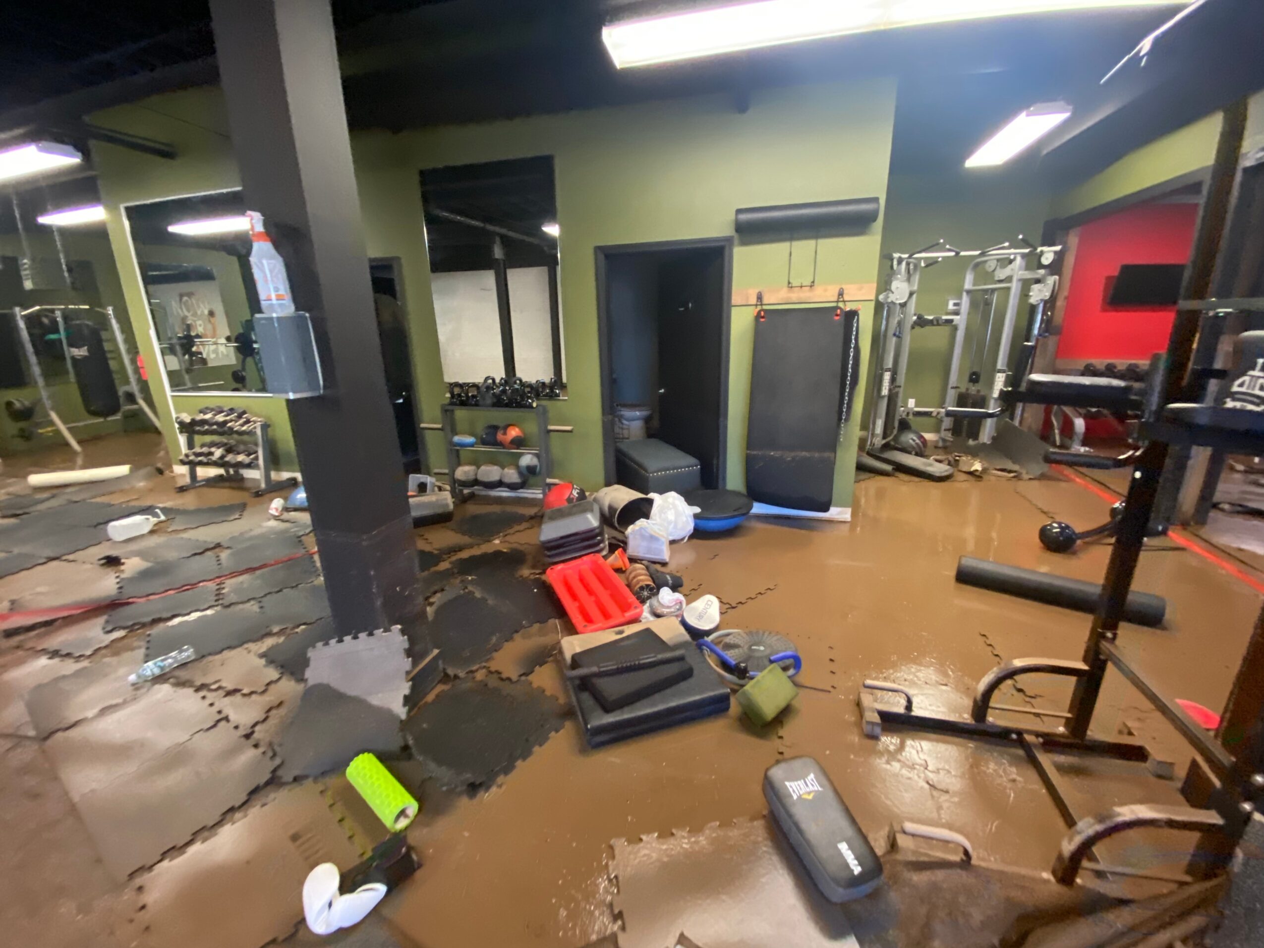 Gym with equipment, mats, and water bottles, showing a cluttered workout area with a brown floor and various fitness gear.