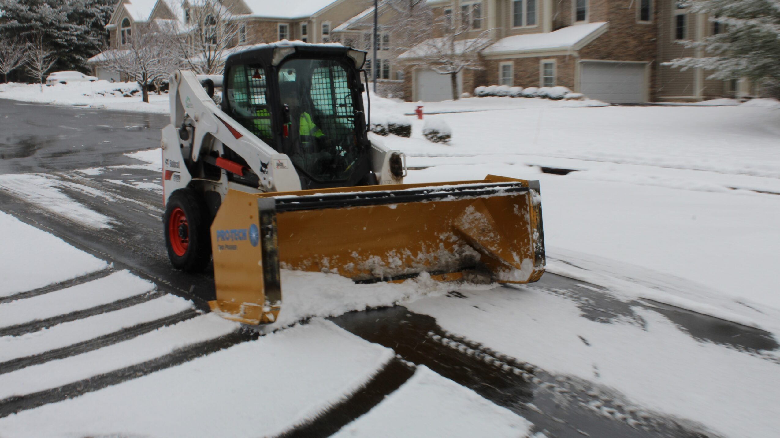 Snow removal machine clearing snow from a residential street, with snow-covered houses in the background.