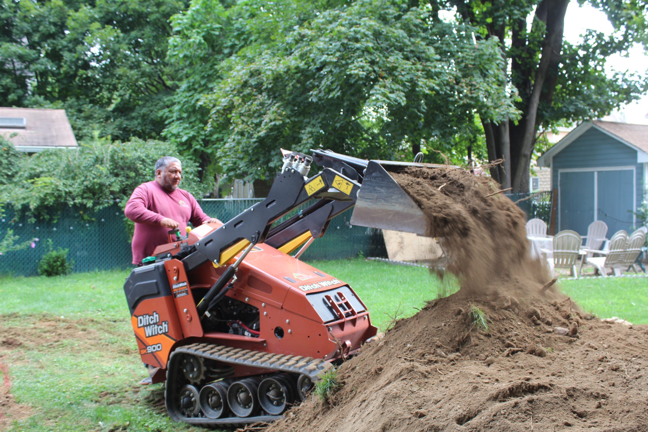 Person operating a small excavator digging and moving dirt outdoors with trees and a house in the background.