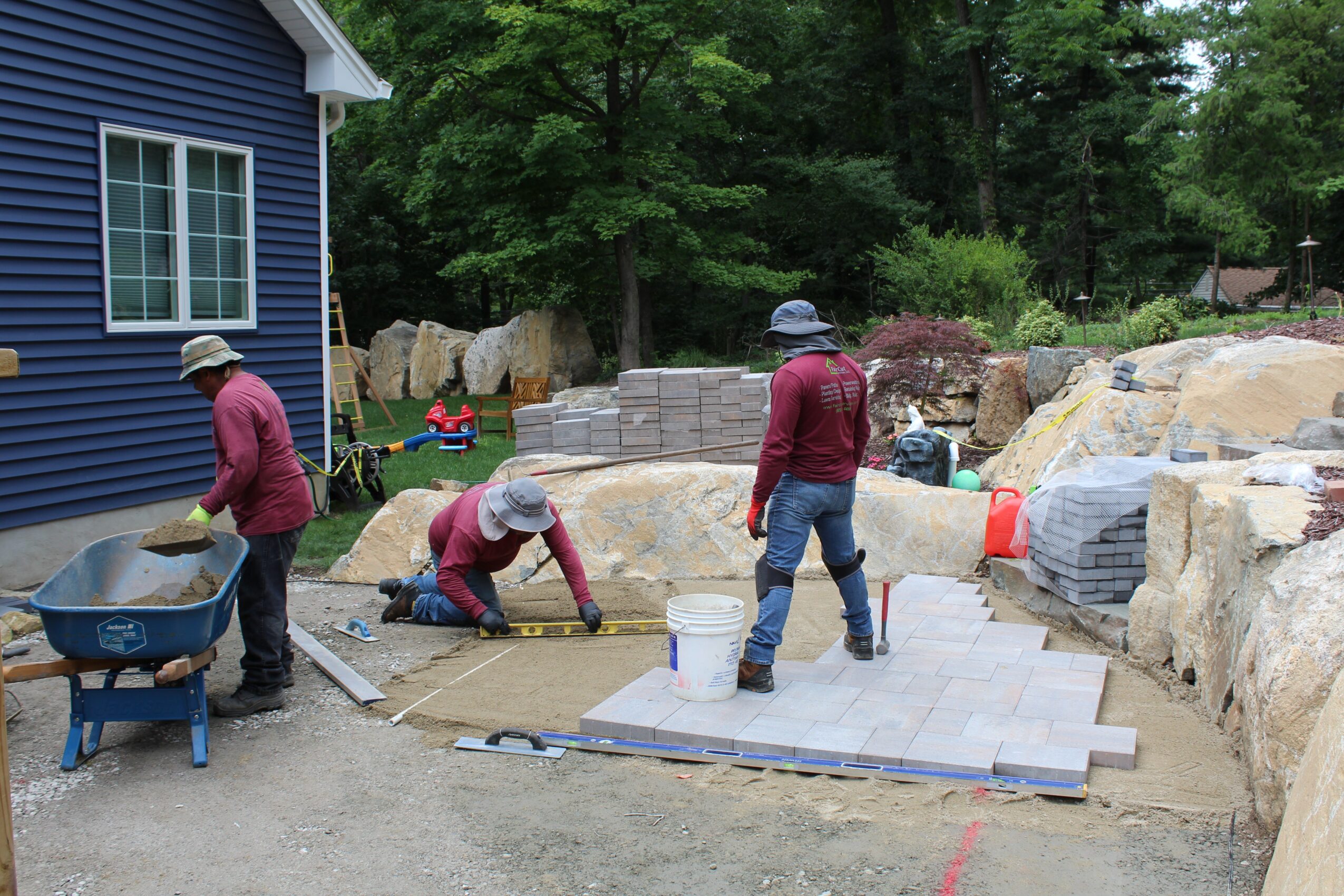 Four people working on a stone patio construction outdoors, with a blue house and trees in the background.