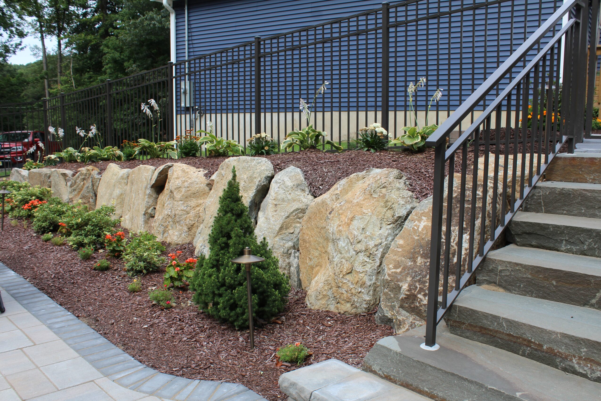 Rock garden with large stones, small shrubs, and flowers along a landscaped area next to stairs and a building.