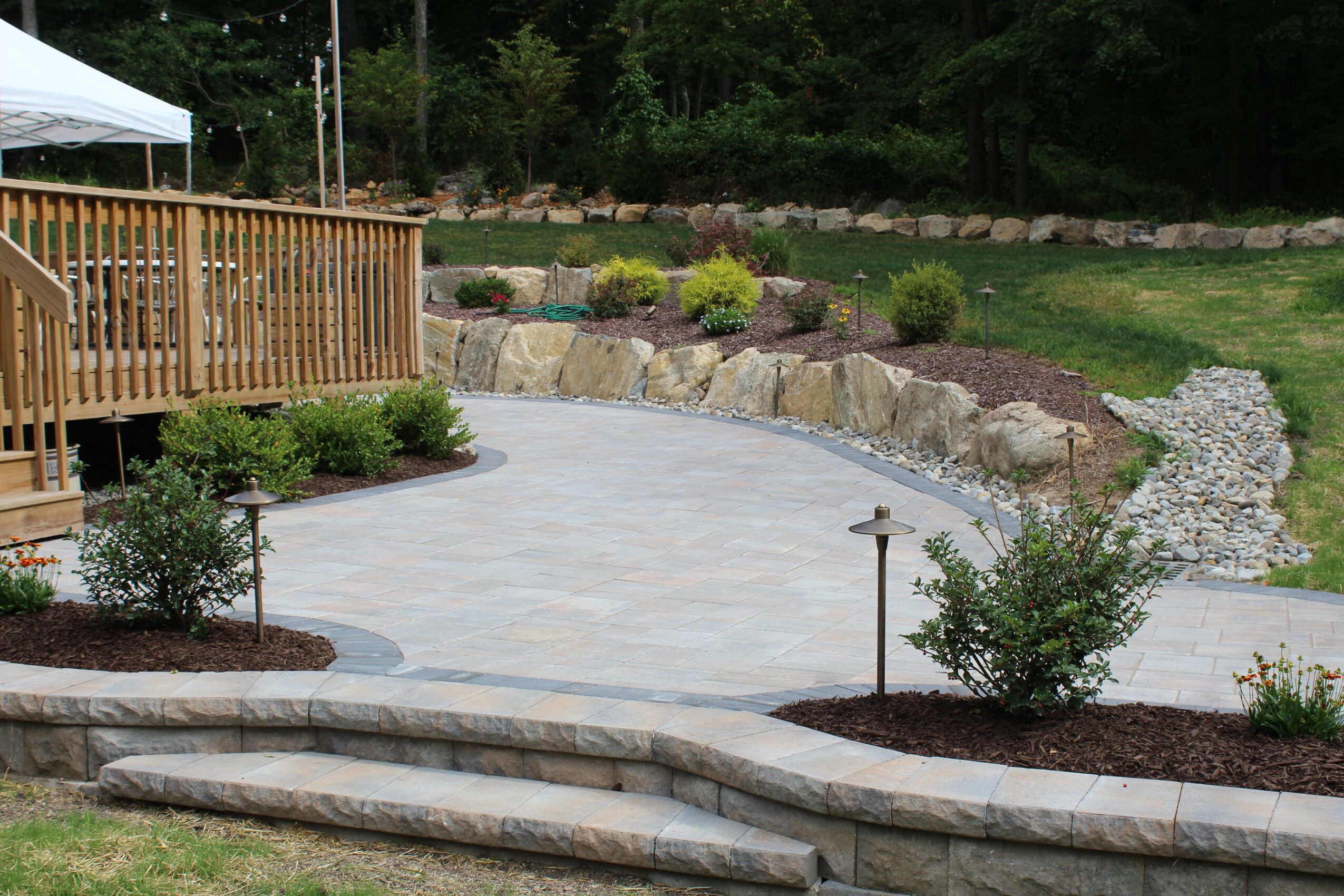 Backyard with wooden deck, paved patio, garden beds, and stone retaining wall, surrounded by trees and greenery.