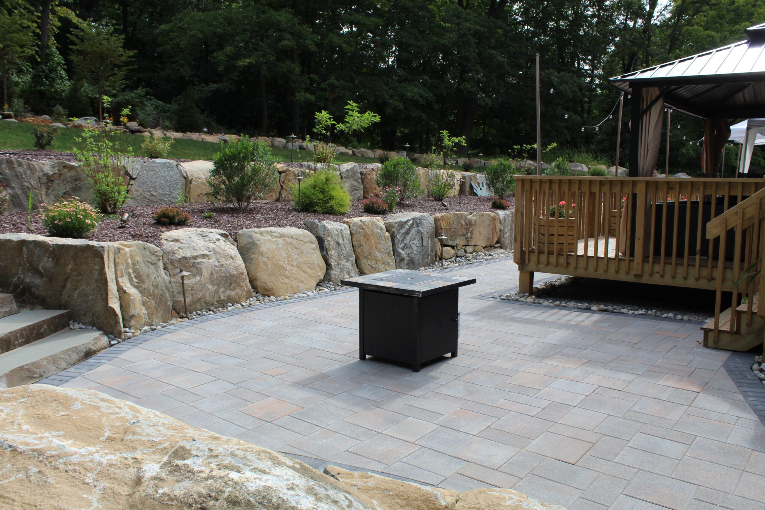 Outdoor patio with stone wall, small table, and wooden deck with stairs, surrounded by trees and plants.