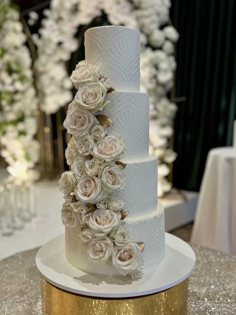 Four-tier white wedding cake decorated with cascading white roses, on a round white stand, with floral background.