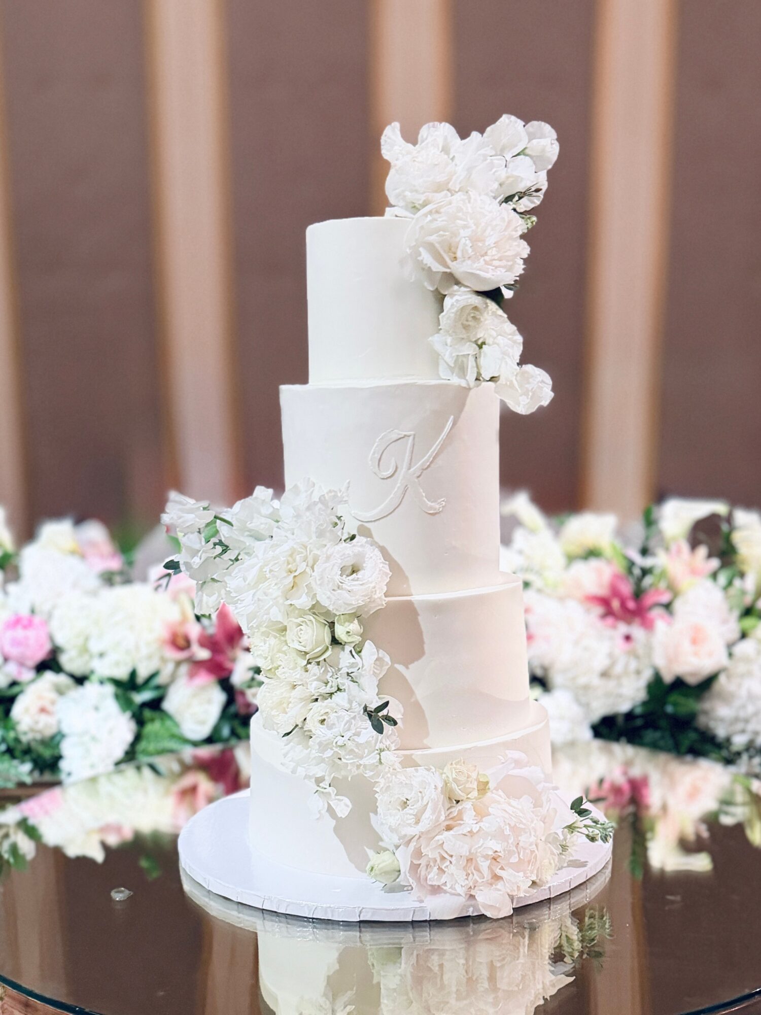 Three-tiered white wedding cake decorated with white and pink flowers, on a reflective surface with floral arrangements in the background.