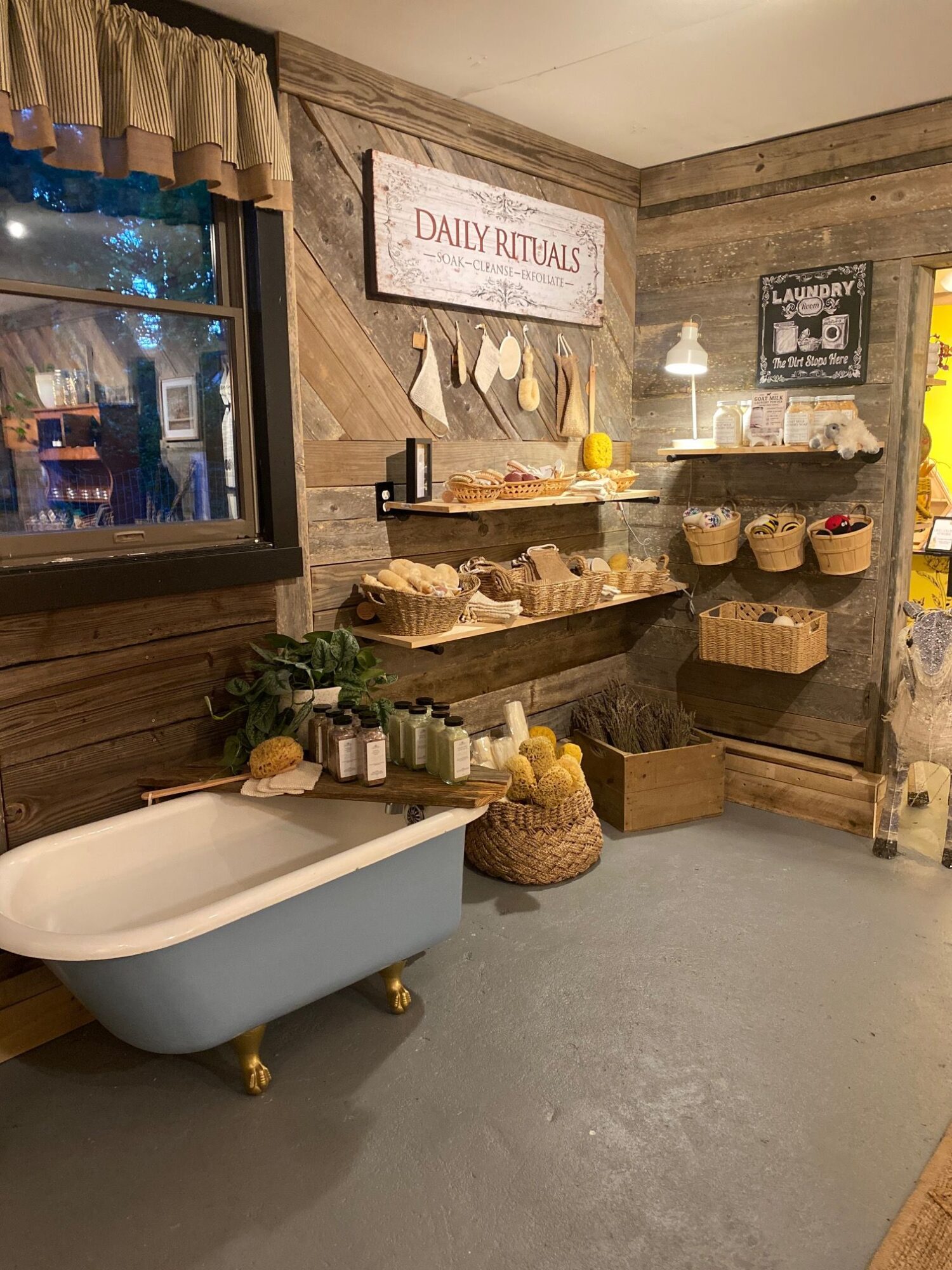Decorative display with a bathtub-shaped container, plants, candles, and small decorative items on a wooden table, with a window and framed art in the background.