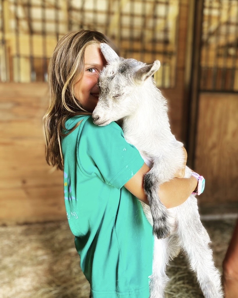 Girl hugging a small goat inside a wooden barn, smiling, with warm lighting.