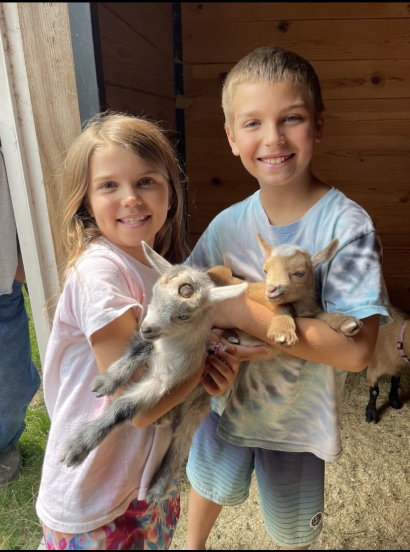 Two children holding baby goats outdoors, smiling, with wooden and fabric background.
